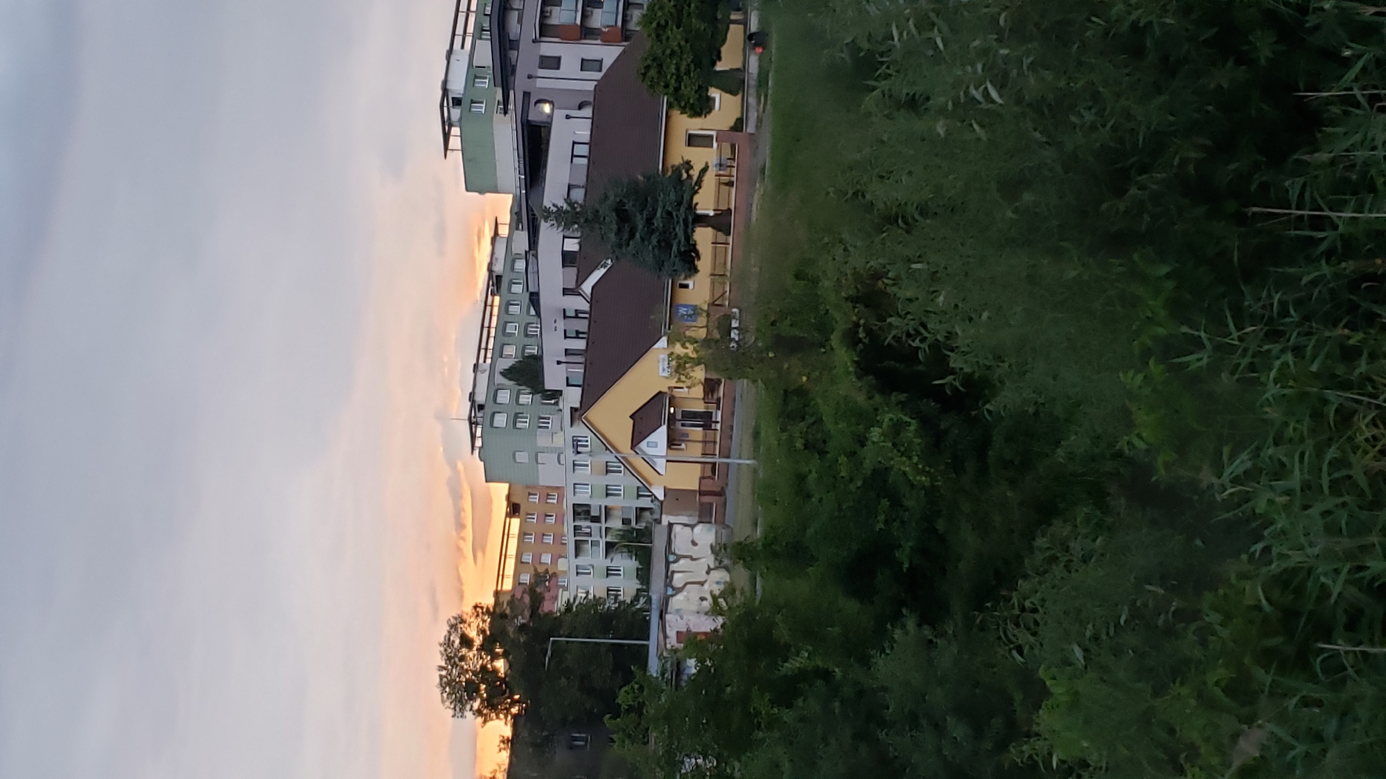 The image depicts a sunset over a residential area in Prešov, Slovakia. In the foreground, dense green foliage partially obscures the view. Behind the foliage, a small, yellow, single-story building with a dark brown roof is visible. This building appears to be a community center or a similar public facility, with a small blue sign and a white door. To the left of the yellow building, a graffiti-covered concrete structure is present.
In the middle ground, several multi-story apartment buildings are situated. These buildings vary in color and design, with some featuring light green facades and dark balconies, while others are predominantly white and grey. The apartment buildings are a mix of modern and older architectural styles.
The background is dominated by a soft, colorful sky as the sun sets, casting an orange and pink glow. The sky is partially covered by light clouds. The overall impression is of a calm, late evening in a suburban or urban environment. The specific location within Prešov is not identifiable from the visual cues alone, but the architectural style of the apartment buildings suggests a typical Eastern European city.