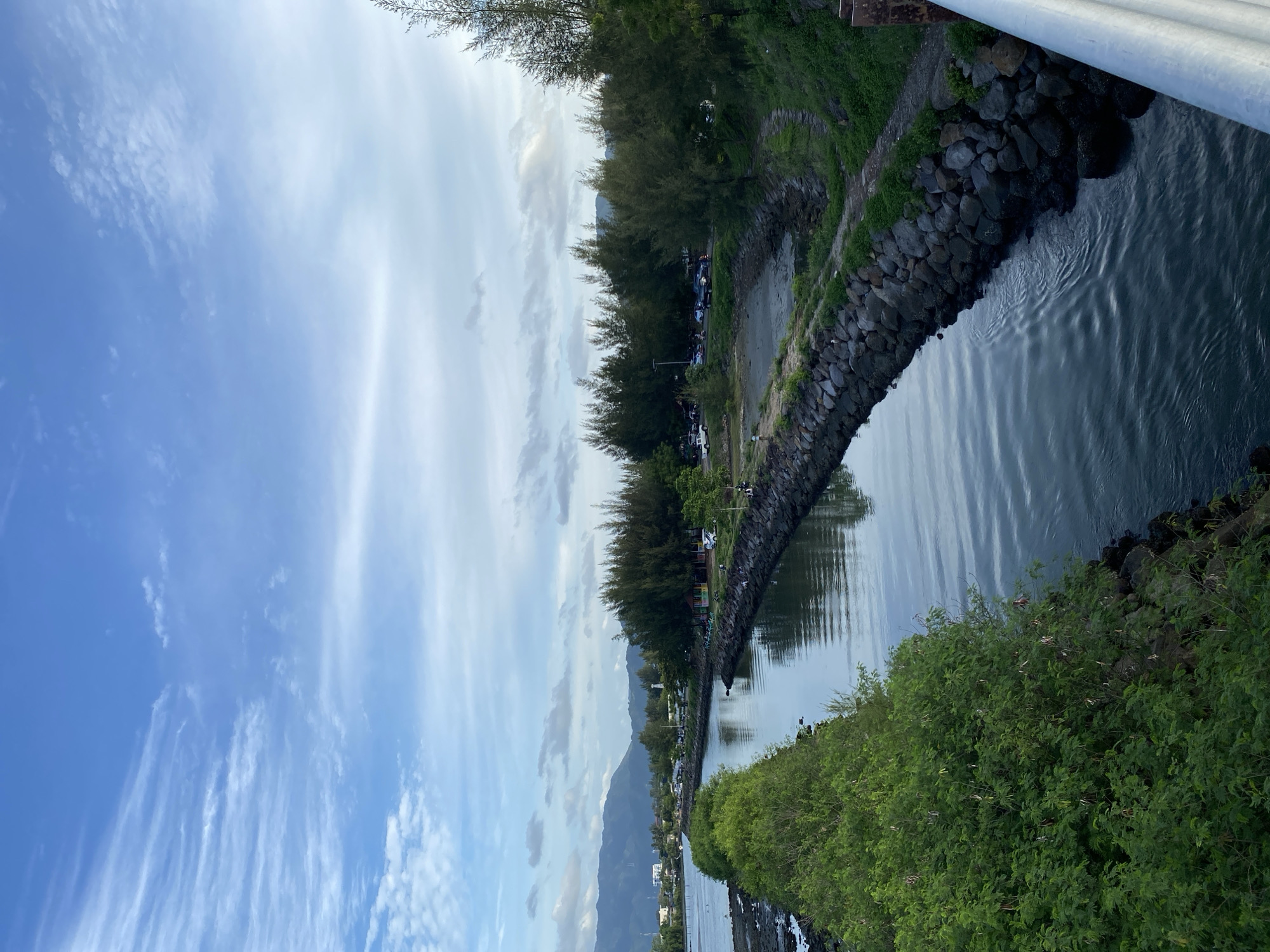 The image shows a canal in Banda Aceh, Indonesia, with a paved path and stone embankment along the right side, and lush green bushes along the left. The canal's water reflects the cloudy sky, which is a mix of blue and white. In the distance, a line of pine trees separates the canal from a road with parked cars and some buildings. Beyond the trees, a mountain range is visible under a hazy sky. A few people are scattered along the embankment, some sitting and others walking. The scene appears to be a public park or recreational area.