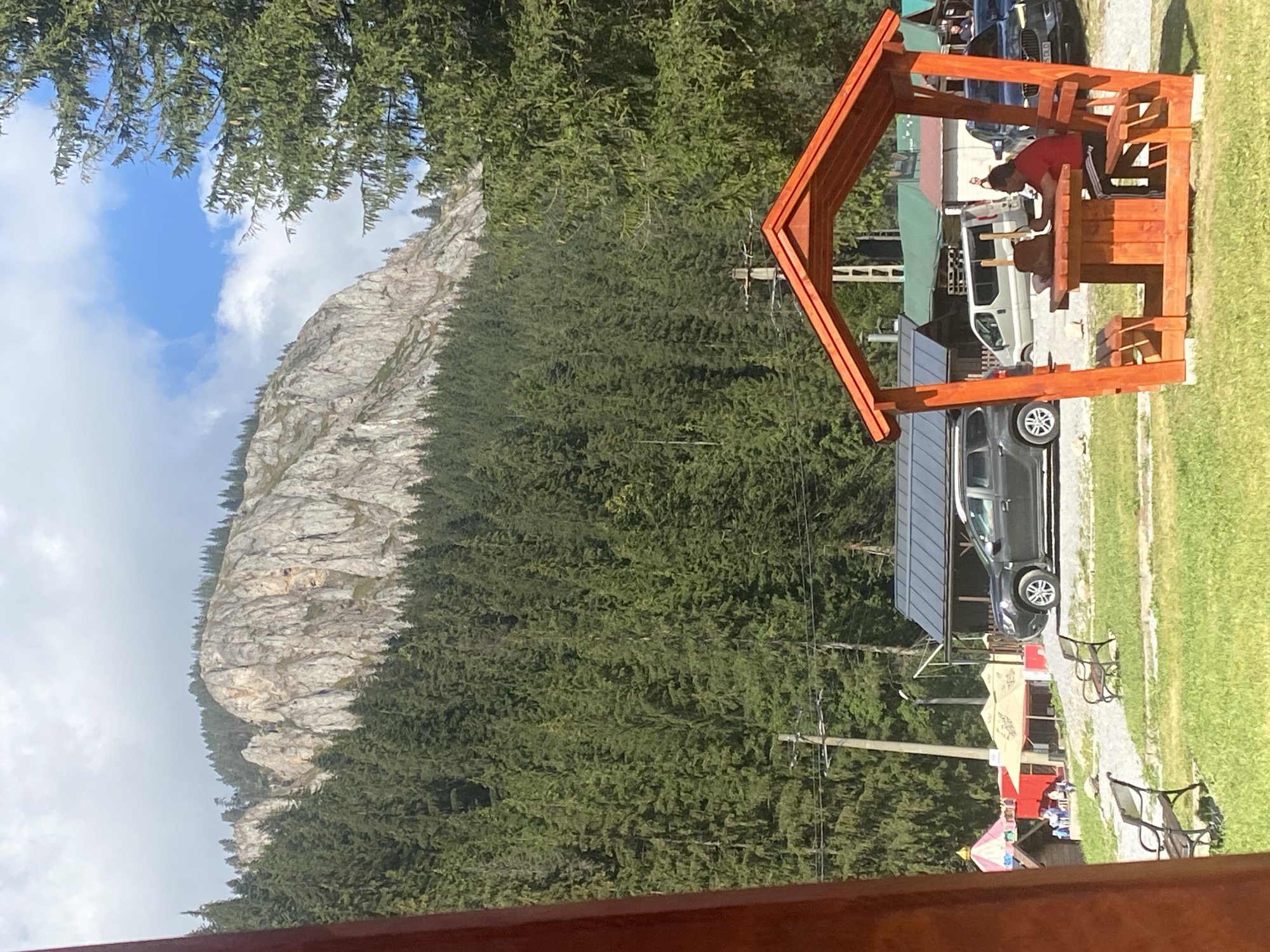 The image captures a scenic view of a mountainous landscape with a dense forest in the foreground. In the lower right portion of the image, a man in a red shirt and black shorts is seated at a wooden picnic table under an orange wooden gazebo. He is facing away from the camera and appears to be engaged in an activity at the table, possibly reading or working, with a brown bag beside him.
Behind the gazebo, there are several vehicles, including a dark-colored SUV and a silver van. Further back, there are buildings with dark roofs, suggesting a village or tourist area. To the left, a small amusement ride or attraction with a colorful tent-like structure is visible, and a few people are scattered around the area. Two empty park benches are situated on the grassy area in the foreground.
The background is dominated by a large, rocky mountain face partially covered by a thick forest of green coniferous trees. The sky above is a mix of blue and white clouds. The scene suggests a day trip or vacation in a mountainous region.
The presence of the amusement ride, picnic area, and parked cars indicates this is a recreational spot. Considering the location specified in Gheorgheni, Romania, this scene likely depicts a tourist attraction or a natural park area within or near the city. The juxtaposition of the natural landscape with man-made structures and recreational facilities is a key characteristic of the scene.