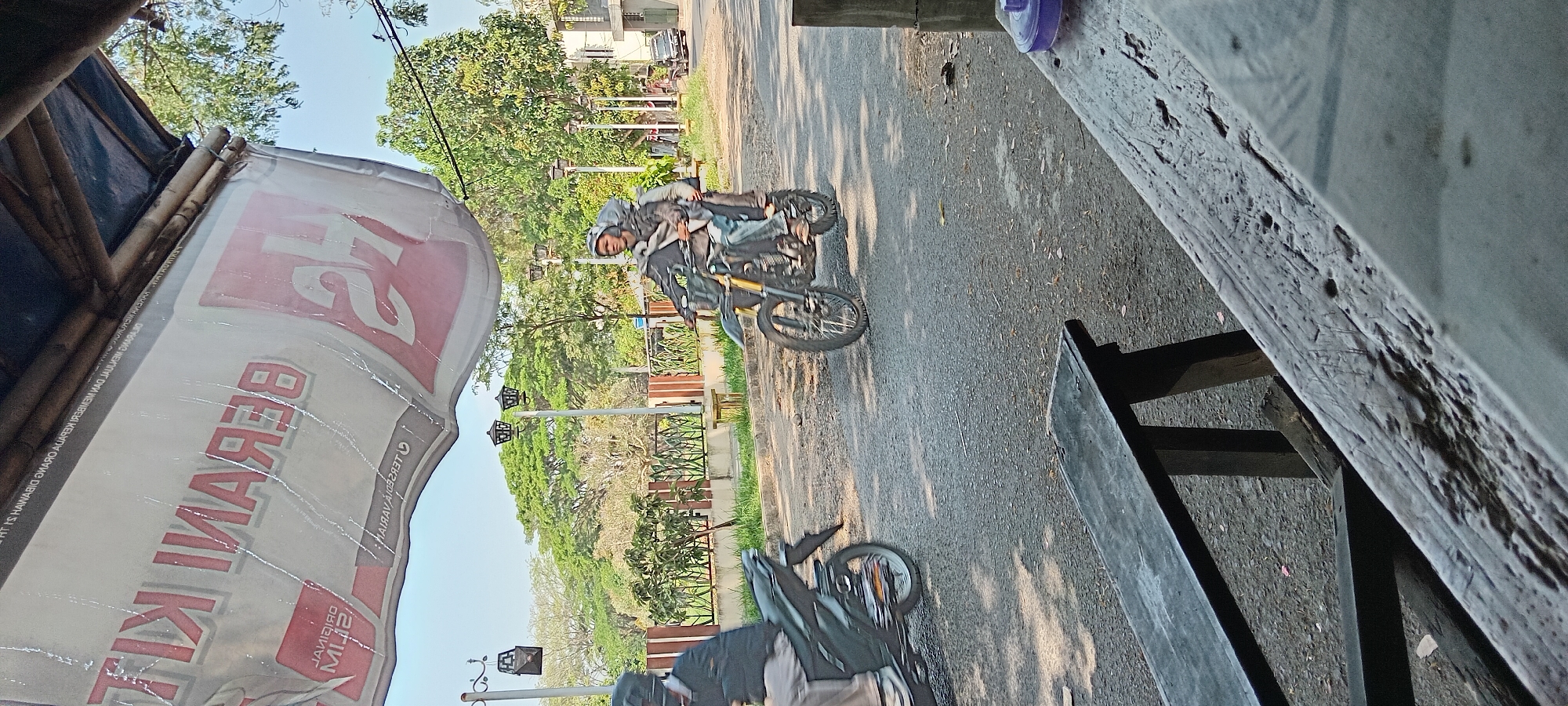 The image depicts an outdoor scene on a sunny day, likely in Tasikmalaya, Indonesia. In the foreground, a rough, textured wooden surface, possibly a table or bench, occupies the lower half of the frame. In the middle ground, a paved road runs horizontally. Two motorcycles are visible on the road. One motorcycle, on the left, is dark and partially obscured, with a rider wearing dark clothing and a helmet. The second motorcycle, a dirt bike with yellow forks, is in the center of the frame, carrying two people. The rider, wearing a helmet and jacket, appears to be looking forward. The passenger behind them is also wearing a helmet and jacket. A banner with red lettering, partially obscured and appearing to be reversed, hangs from a structure above, casting a shadow. Beyond the road, a manicured area with grass, trees, and decorative lamp posts is visible. In the background, more trees and buildings can be seen under a clear sky.
