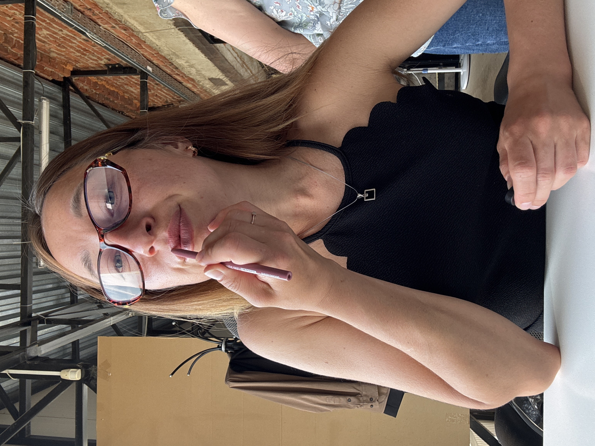 A woman with brown hair and glasses applies lip liner. She wears a black top and a silver necklace. The background suggests an urban setting, potentially in Chaikovsky, Russia. The scene is framed by architectural elements of a city building.