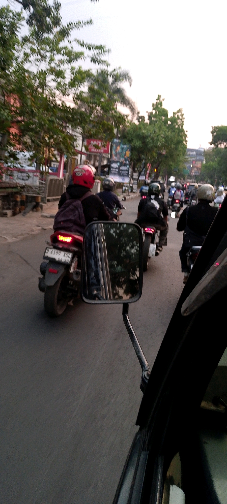 The image shows a street scene in Bandung City, Indonesia, with a line of motorcycles and riders on a road. In the foreground, a motorcycle with a red helmet and backpack is visible, with its license plate reading "D 4199 XGL". A rearview mirror of another vehicle is positioned to the right, reflecting trees and the sky. Further down the road, several other motorcycles and riders are seen, with some traffic and buildings lining the street. The sky is overcast, suggesting it may be late afternoon or early morning. The overall impression is that of a typical urban street environment with active motorcycle traffic.