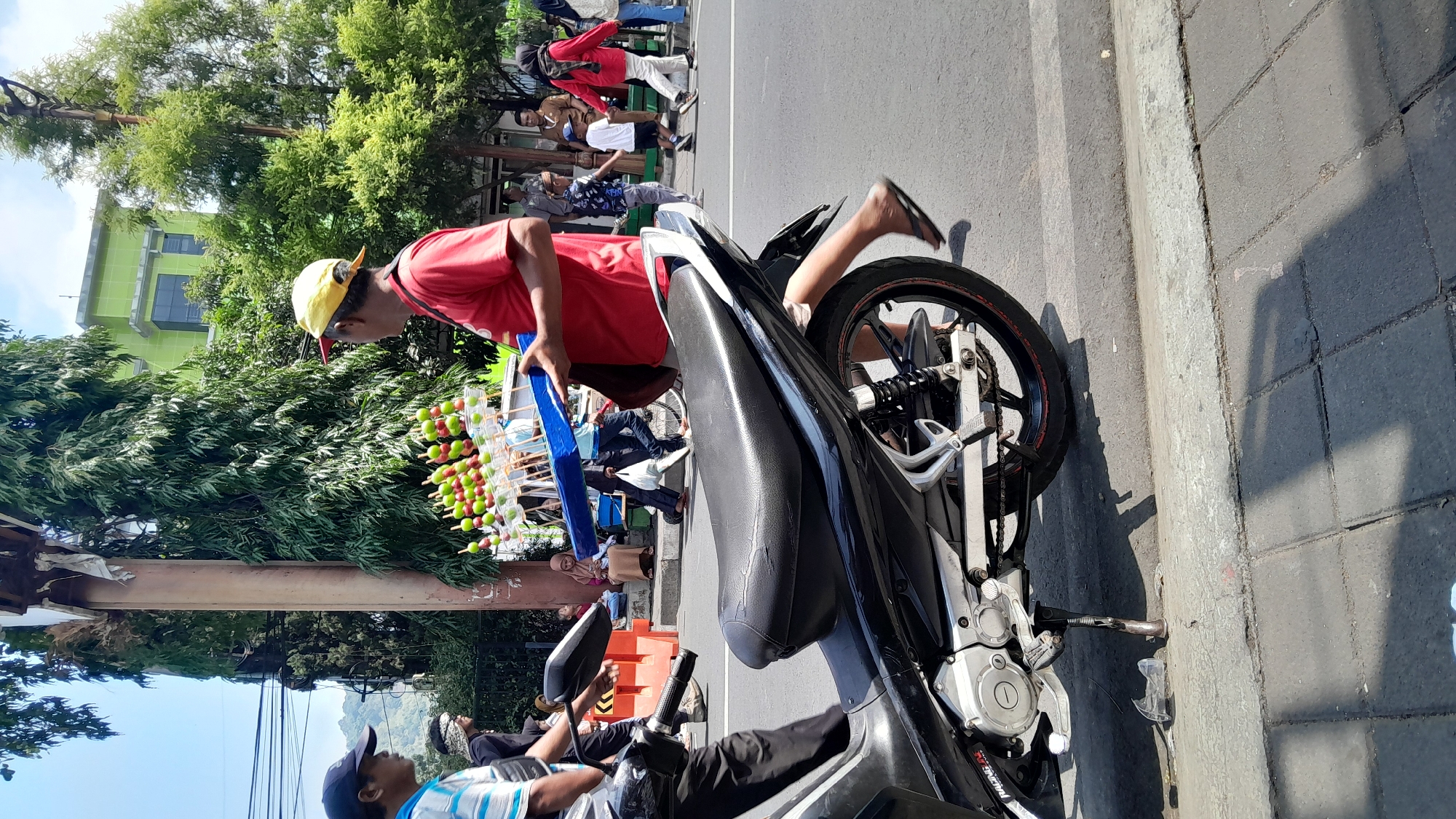 In Sumedang, Indonesia, a street vendor wearing a red shirt and a yellow cap holds a blue tray filled with fruit skewers. He stands on a sidewalk adjacent to a busy street, with a black motorcycle parked in the foreground. The vendor appears to be interacting with another person, possibly a customer, who is partially obscured by the motorcycle. Other pedestrians are visible in the background, along with trees and buildings. The scene suggests a typical urban environment in Indonesia, with a focus on street commerce and daily life.