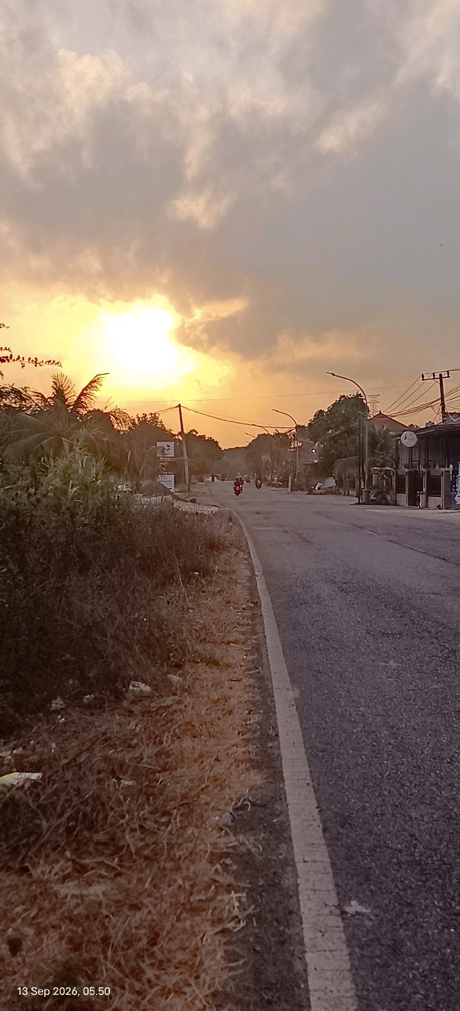 An image taken in Mojopetung, Indonesia, captures a roadside scene during what appears to be sunset or sunrise. The sky is filled with dramatic clouds, some illuminated by the sun's bright glow on the left side of the frame, creating a warm, orange-yellow hue. A road stretches into the distance, with a white line marking the edge of the lane. On the left side of the road, there is a patch of dry, overgrown vegetation and some scattered debris. Further down the road, several motorcycles are visible, suggesting traffic. Buildings and trees line the right side of the road, with utility poles and wires also present. The overall atmosphere is somewhat hazy, typical of tropical weather conditions. A timestamp at the bottom left corner of the image reads "13 Sep 2026, 05.50."