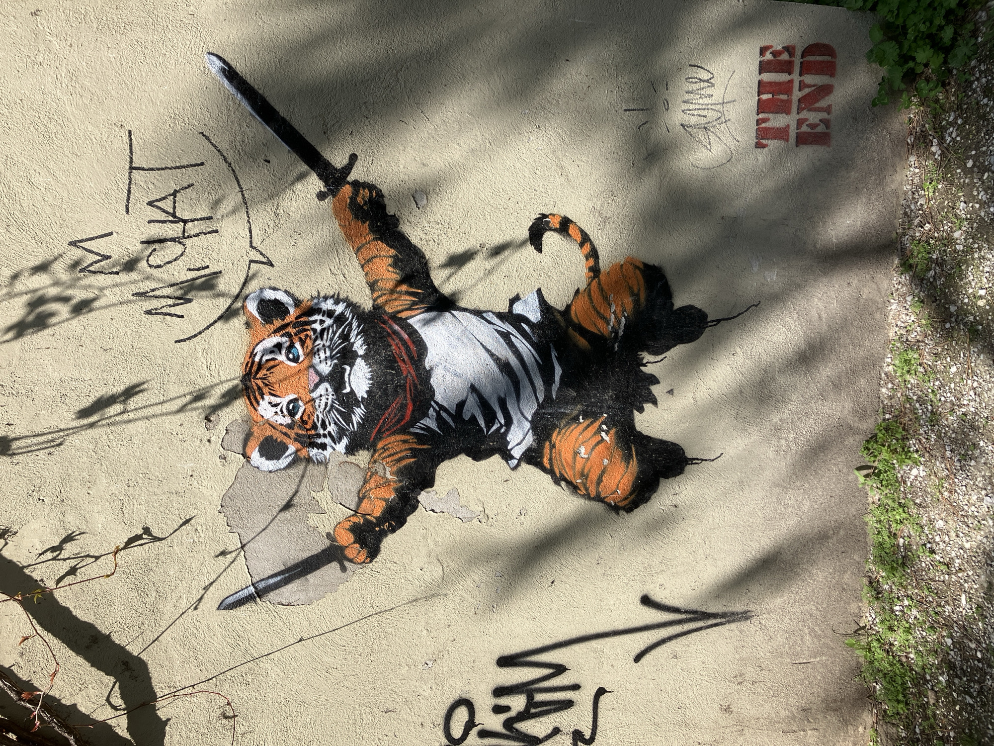 The image shows a street art mural on a light yellow concrete wall. The mural features a tiger lying on its back, holding two swords. The tiger has orange and black stripes, a white belly, and blue eyes. Above the tiger on the left, there is graffiti that reads "WHAT". Above the tiger on the right, there is graffiti that reads "THE END". Below the tiger, there is an arrow and more indecipherable graffiti. To the right, there is some foliage growing on the wall. Some shadows are visible across the wall. The location of the artwork is in the city of Unknown, Unknown.