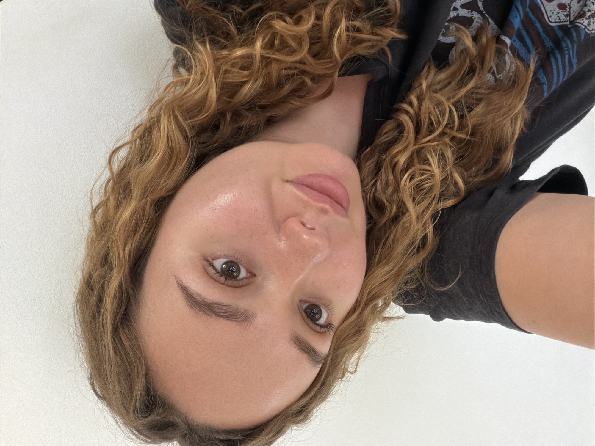 A portrait-style image, rotated 90 degrees counter-clockwise, depicts a young woman with light brown, curly hair looking directly at the camera. She has brown eyes, well-defined eyebrows, and light pink lips. Her skin appears clear with a slight natural sheen. She is wearing a dark-colored t-shirt, possibly black or dark grey, which features a faint blue and white graphic design visible over her right shoulder. The background is a plain, light-colored wall, suggesting an indoor setting. The overall expression is neutral. This scene is located in Ramat Gan, Israel. There is no visible text in the image.