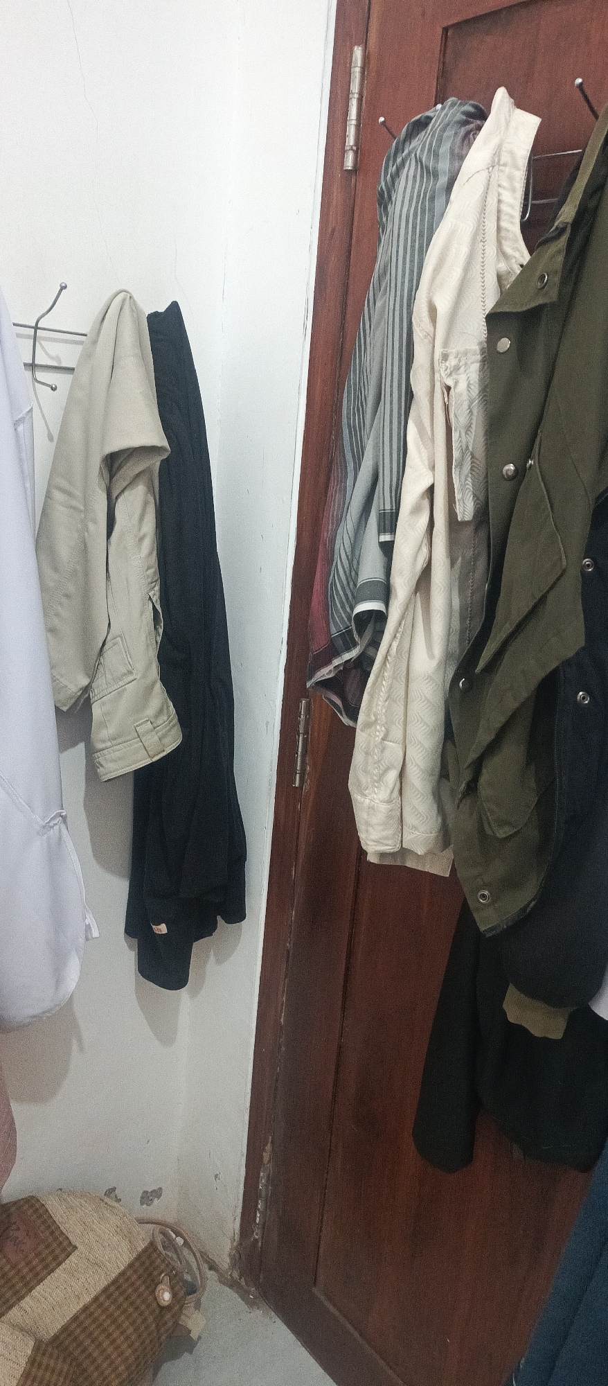 A medium shot captures a section of a wall with a dark brown wooden door on the right. Several items of clothing are hanging on hooks or the door frame. On the left side, against the white wall, a tan jacket and a black garment are hung on metal hooks. Partially visible to the left of the tan jacket is a white garment. On the right side, hanging on the door frame, are multiple garments: a striped shirt, a cream-colored shirt with a textured pattern, a dark olive green jacket, and a black garment. The floor in the lower left corner shows a woven bag with brown and tan checkered patterns. The environment appears to be an indoor domestic space, possibly a bedroom or hallway, in Arjasa, Indonesia.