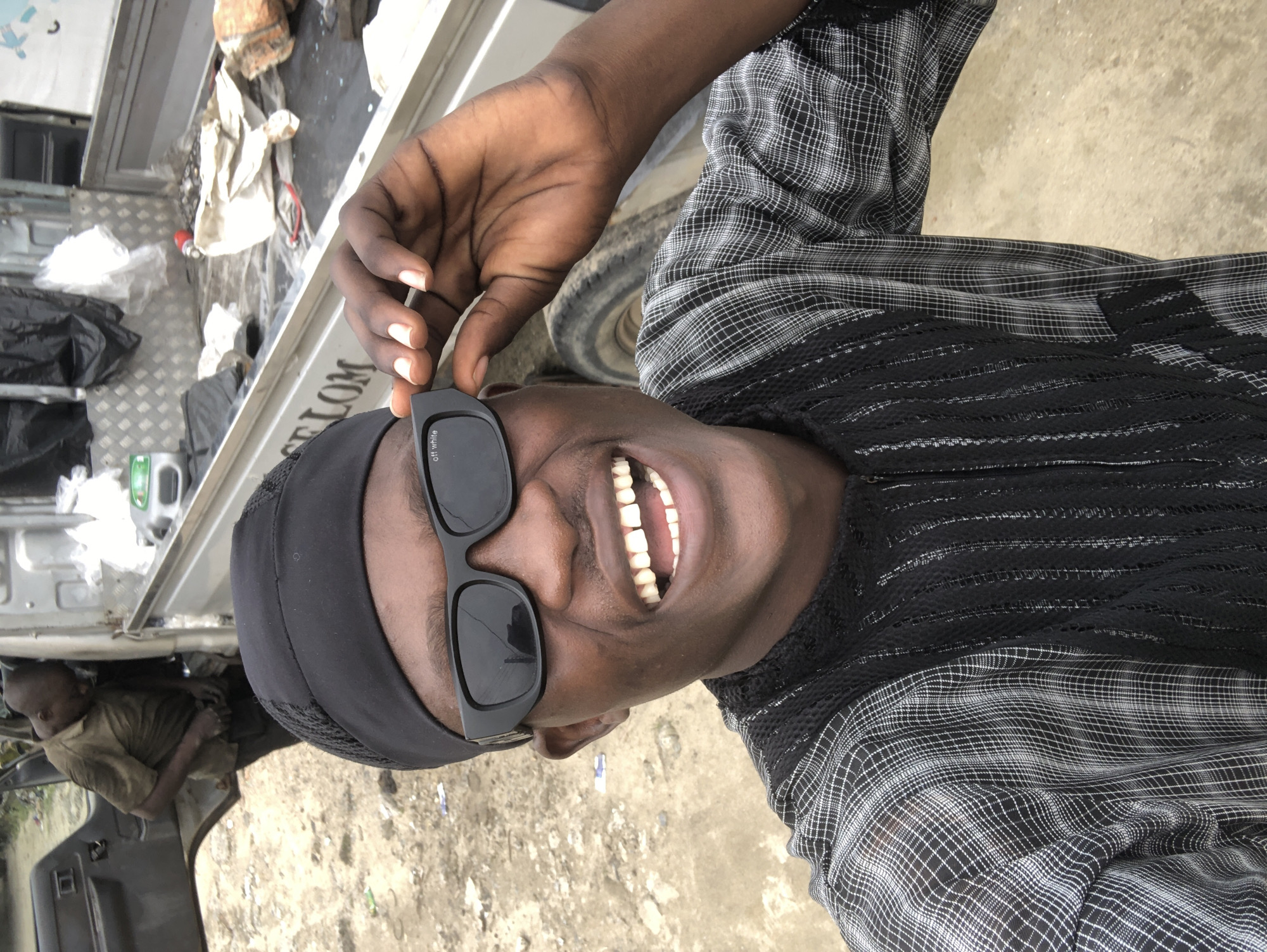 A medium shot shows a man in the foreground wearing sunglasses and a black bandana. He is smiling broadly, revealing his teeth. His hand is raised to adjust the sunglasses. He is wearing a black and white checkered shirt with black detailing on the chest. In the background, an open truck is visible, loaded with various items including black trash bags and white plastic sheeting. Another man, wearing a brown t-shirt and dark pants, is partially visible inside the truck's cab, facing away from the camera. The ground is dirt and appears somewhat uneven. The setting is an outdoor, possibly industrial or service area, likely in Ado, Nigeria, given the context. The lighting is bright, suggesting daylight.