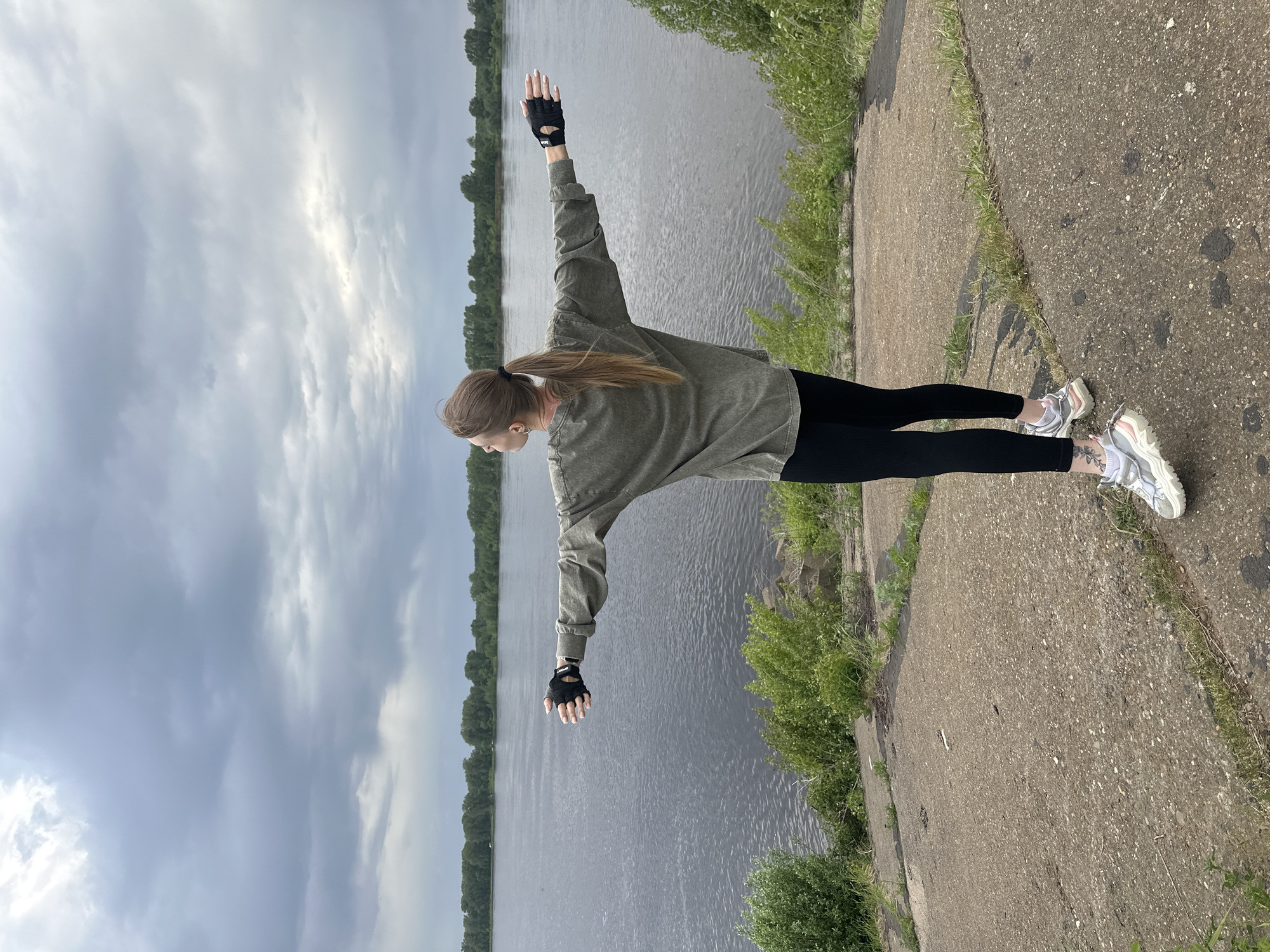 A woman stands on a concrete path in Chaikovsky, Russia. She is wearing black leggings, a green long-sleeved shirt, fingerless gloves, and white sneakers. Her arms are outstretched, forming a "T" shape. A river is visible behind her, flanked by trees. The sky is overcast. The path is cracked in places and has grass growing on its edges.