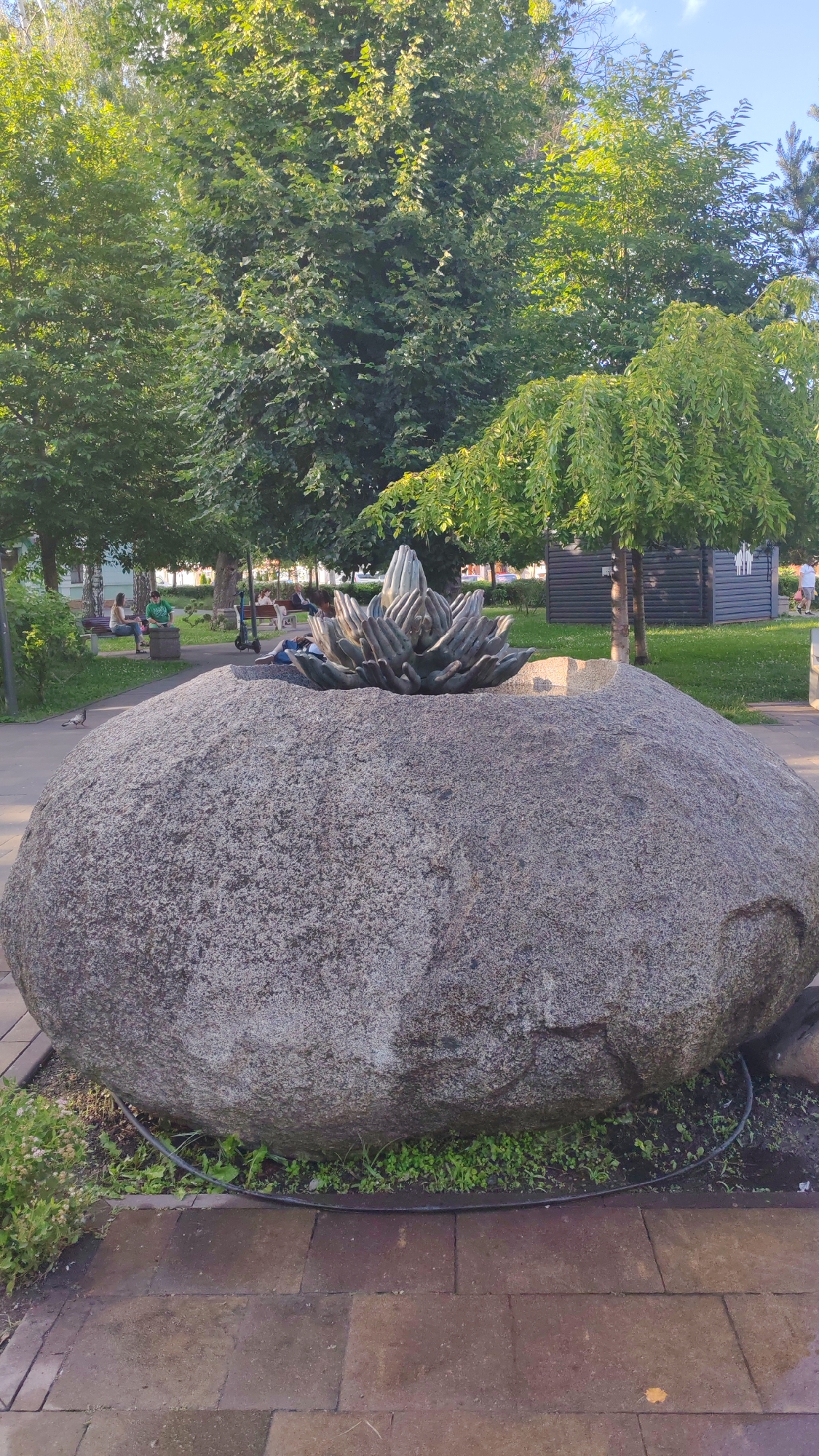 In the city of Unknown, Unknown, a large, grey, boulder-like sculpture featuring stylized hands forming a lotus-like shape sits atop a massive rough-hewn granite boulder. The sculpture is centrally located in a park-like setting with lush green trees providing a dappled canopy. Paved pathways and grassy areas surround the boulder. In the background, people are visible seated on benches, engaged in conversation or relaxing. Some individuals are lying down near the sculpture. Additionally, a pigeon is seen walking on the pathway in the foreground. The scene is bathed in natural light, suggesting it is daytime.