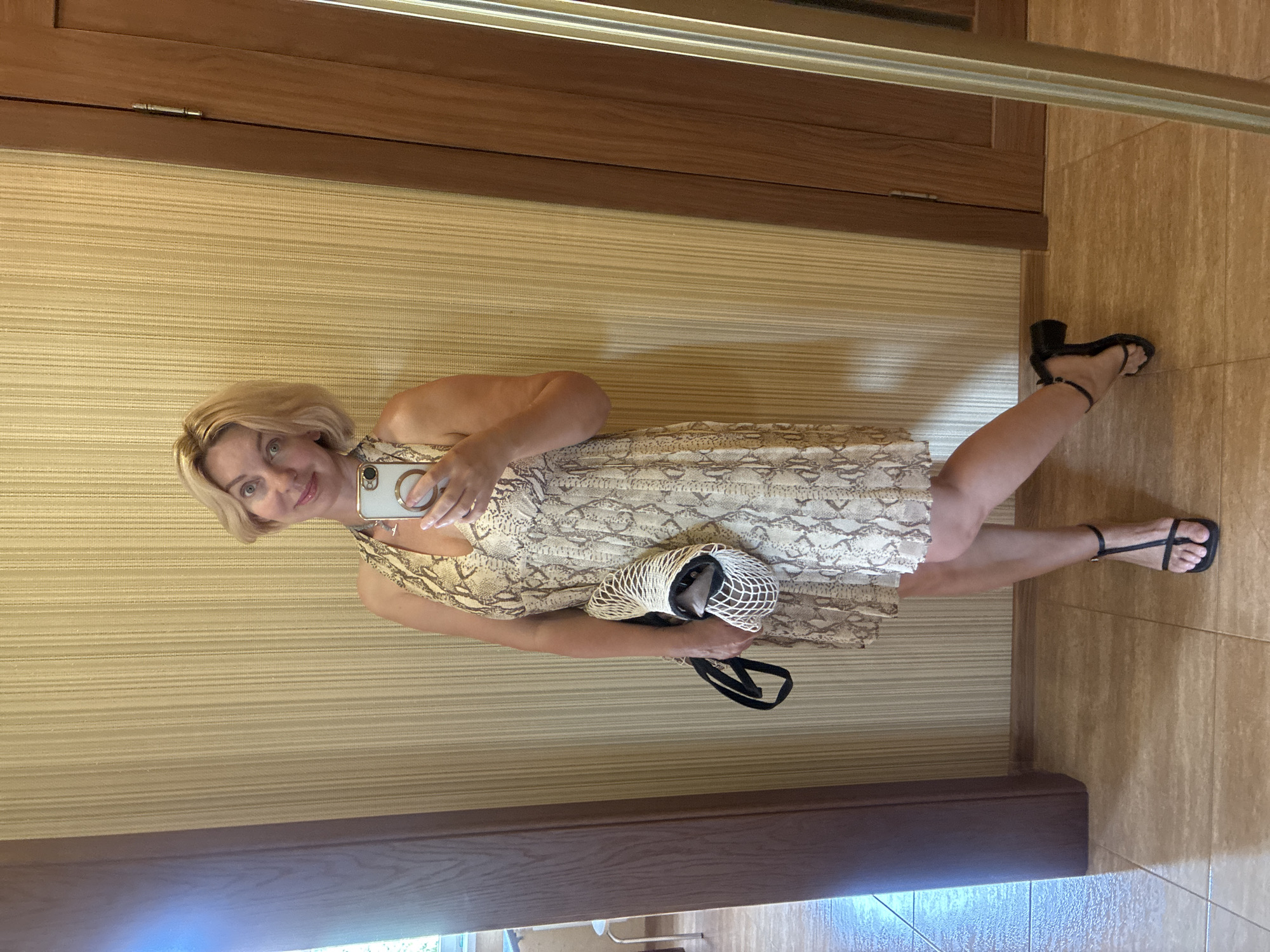 In an indoor setting, a blonde woman with short hair is taking a mirror selfie. She is wearing a beige snakeskin print halter dress and black strappy sandals with block heels. She is holding a white net bag containing an item, possibly a bottle, and a black purse strap. The woman is standing in front of a textured beige wall with vertical stripes. To her left is a dark wood door frame and a reflection of part of a room, including a window. To her right is another dark wood door. The floor is tiled in a light brown and beige pattern. The scene appears to be in a residential interior.