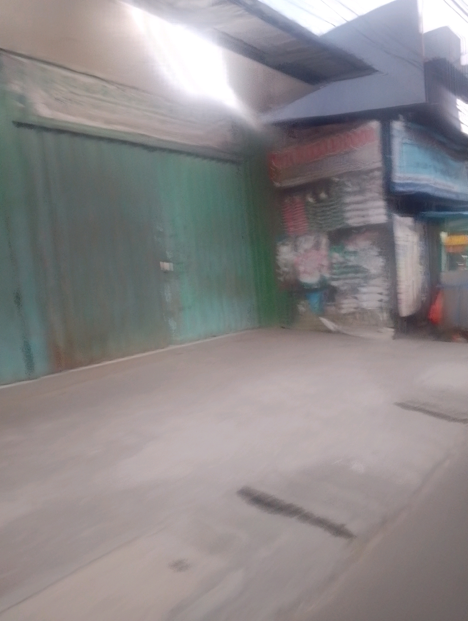 The image depicts an outdoor scene in Depok, Indonesia, likely in front of a commercial establishment. In the foreground, a light-colored concrete surface, possibly a sidewalk or parking area, shows signs of wear and some dark smudges. A white line is visible near the bottom edge, marking a boundary or separation.
Dominating the mid-ground is a large, faded green metal gate or garage door, which appears to be slightly weathered. To the right of the gate, a building with a corrugated metal facade is visible. This building features several signs, some with what appear to be colorful graphics or text, though they are mostly indistinct due to the image's blurriness and low resolution. Above the buildings, a part of a roof or awning structure is visible, with bright light reflecting off its surface.
The overall scene is characterized by a muted color palette, with greens, grays, and light browns. The lighting suggests it is daytime, with a bright light source from above, possibly the sun, creating reflections and shadows. The image is generally out of focus, making specific details difficult to ascertain. There are no people clearly visible in the scene.