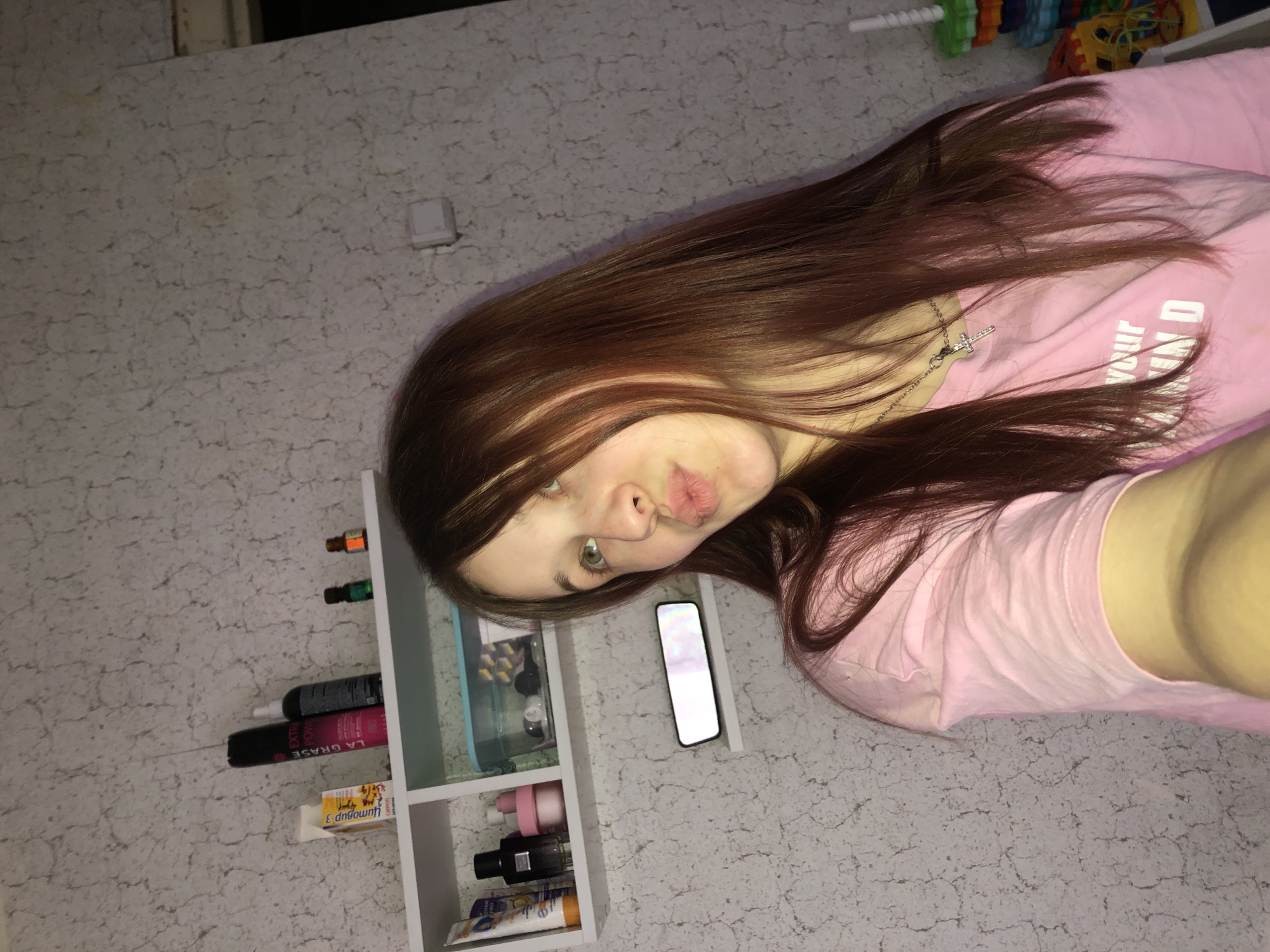 The image depicts a young female, approximately 15-25 years old, with long, straight, reddish-brown hair, and light-colored eyes. She is positioned slightly to the right of the frame, looking upwards and to the left, with her lips puckered in a "duck face" expression. She wears a light pink t-shirt with partial white text visible. A silver chain with a small silver cross pendant hangs around her neck. Her right arm is extended, suggesting she is taking a selfie.
The background consists of an indoor wall covered in light-colored, textured wallpaper featuring a mottled, crack-like pattern. A white multi-compartment shelf is mounted on the wall. On the upper tier of the shelf, from left to right, are a yellow and white box labeled "Цитовир-3", a black aerosol can labeled "LA GRASE EXTRA POWER", a dark spray bottle, a small dark green dropper bottle, and a small amber dropper bottle. On the lower tier of the shelf, from left to right, are a white and orange tube with Cyrillic text, a dark bottle, a pink jar, and a translucent light blue plastic container holding assorted small items. Below the shelf, a small, rectangular, horizontally oriented device with an illuminated, colorful display is affixed to the wall. To the far right, a stack of colorful children's toys is partially visible. A white square object, possibly an electrical outlet or light switch, is also visible on the wall further right. The specific location within the city of Фоки, Russia, cannot be determined from the provided image.