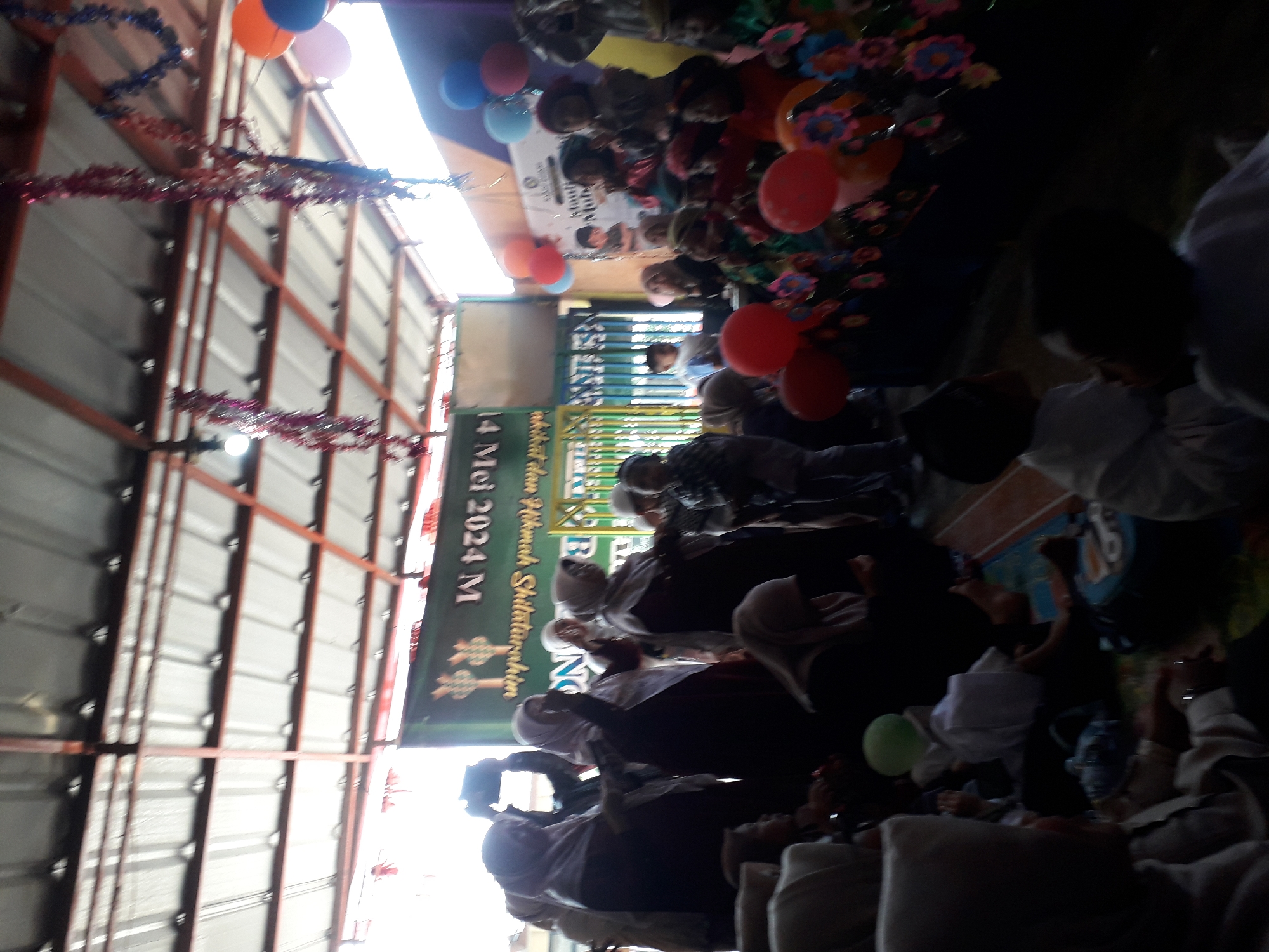 The image captures an outdoor gathering in Cankuang, Indonesia, likely a school event or celebration. The scene is crowded with people, mostly children and women, dressed in traditional Islamic attire, including hijabs and peci hats. Some individuals are holding or taking photos with their phones. Colorful decorations, such as tinsel garlands and balloons in red, orange, pink, and blue, adorn the area, suggesting a festive occasion. A banner with Arabic writing, partially visible and inverted, indicates a date of "14 Mei 2024 M" and mentions "Madrasah Ibtidaiyah Shaltunirashim". The event appears to be taking place under a covered structure with a metal roof supported by beams. Children are seated on mats and the floor, engaged in activities or observing the proceedings. The overall atmosphere seems joyful and community-oriented.