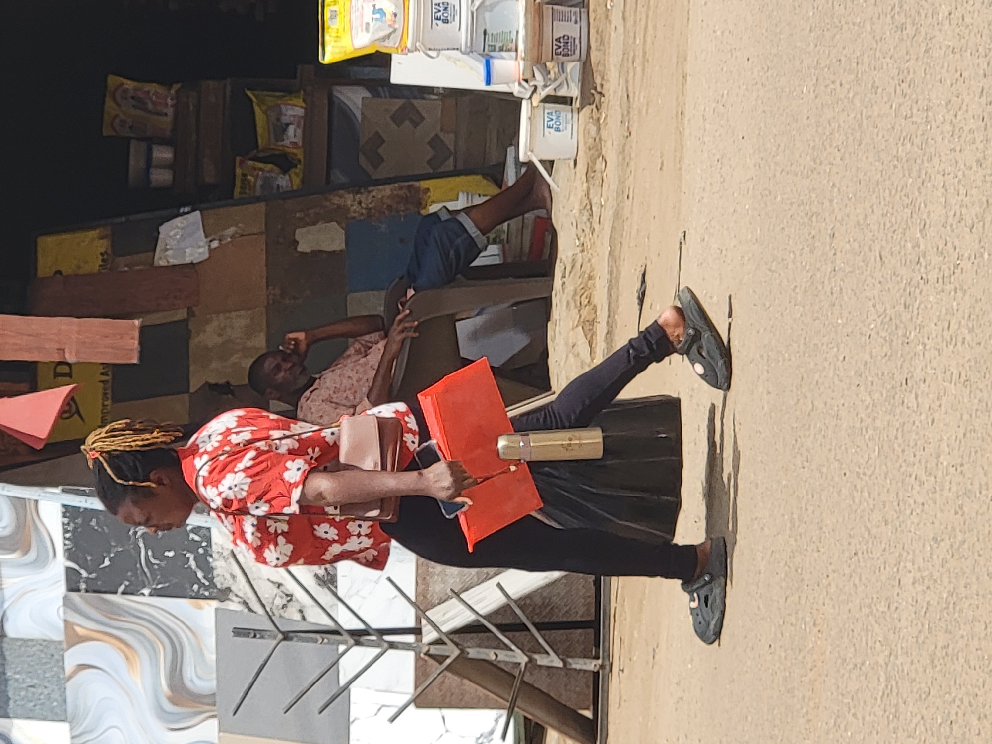 A woman with blond dreadlocks, wearing a red floral shirt and black leggings with gray crocs, walks down a street in Maiburiji, Nigeria. She carries a tan purse and an orange package/folder. Behind her, a man lies on a bench, smoking a cigarette. In the background, there's a store selling boxes of EVA BOND. The pavement appears to be concrete, and the woman appears to be walking on the sidewalk. Tiles are displayed near the woman.