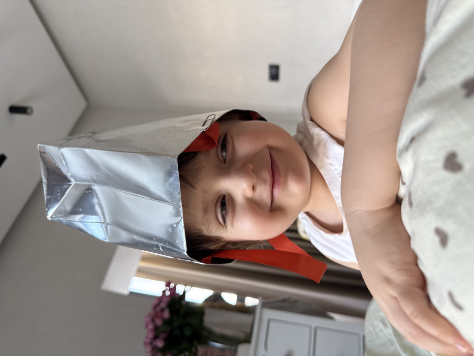 A young child, approximately two to four years old, is depicted in a medium close-up, smiling with their head slightly tilted to the viewer's right. The child wears a white tank top and a conical, crudely constructed hat made of reflective silver foil, secured by red ribbon straps, one of which appears to be fastened with a paperclip. Their forearms rest on a light-colored bedding item patterned with small, dark heart shapes.
In the blurred background to the left, a white multi-drawer bedside table is visible, topped with a clear vase containing pink flowers. Behind this, a window with light-colored curtains allows bright daylight into the room. The background to the right consists of a plain, light-colored wall. The ceiling, also light-colored, features two black, cylindrical spotlight fixtures. The specific location in the city of Khimki, Russia, is not discernible from the visual information provided in the image.