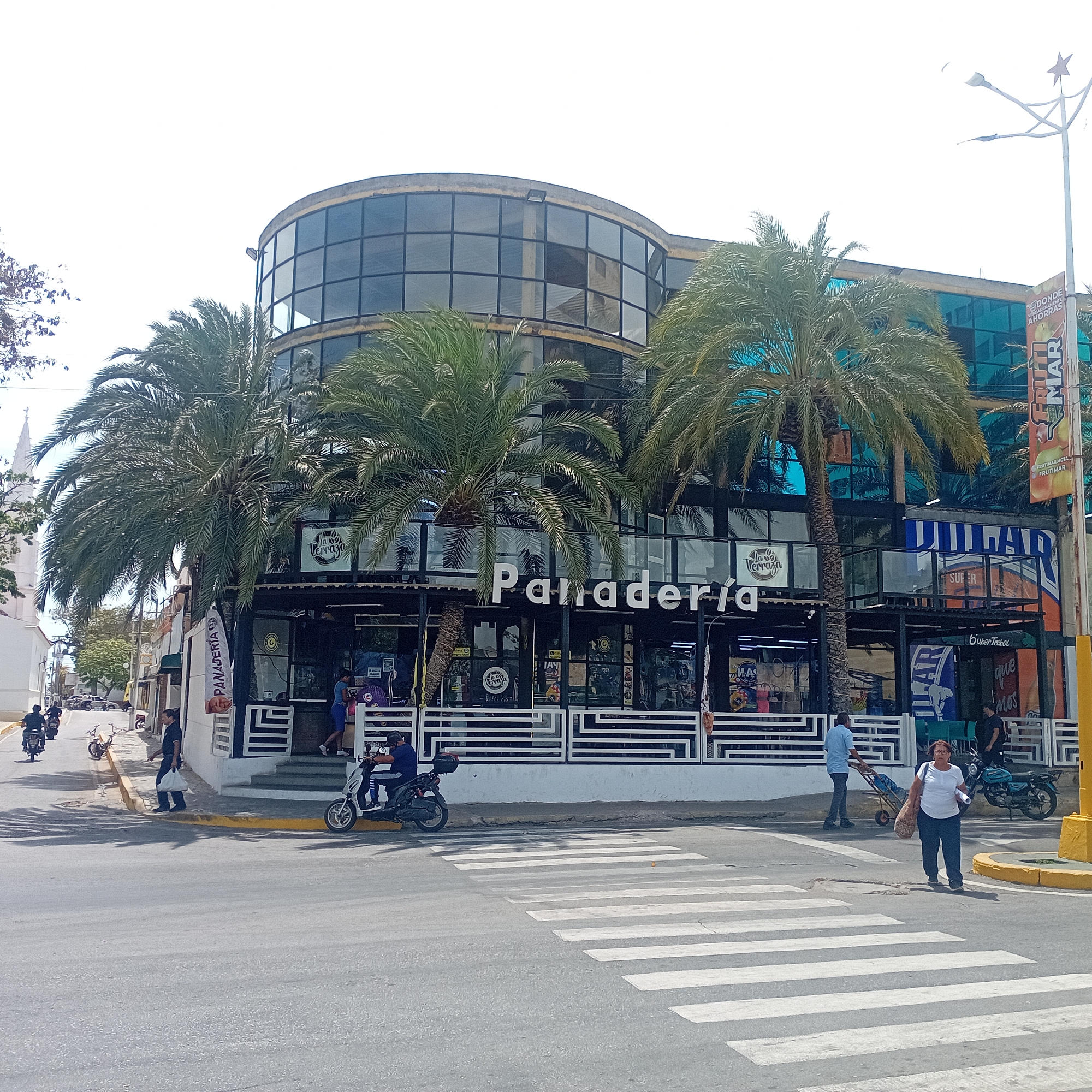 The image depicts a street scene in an urban location. A building stands prominently, featuring a bakery called "Panaderia" on the ground floor. The building has a curved, glass-walled upper section. Several palm trees are visible in front of the building. Pedestrians and motorcycles are present on the street. A woman is walking across a zebra crossing. A man is walking on the sidewalk, pulling a cart. A church steeple can be seen in the background. The environment appears to be sunny.