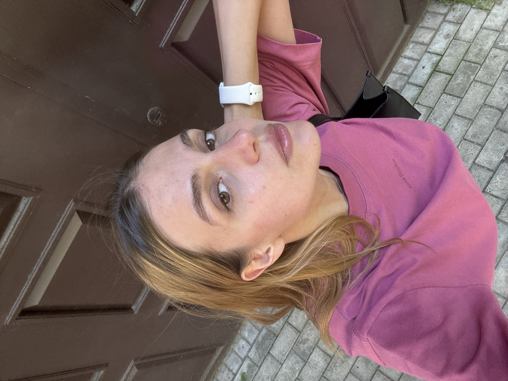 A woman is positioned at an unconventional angle within the frame, appearing sideways. She is wearing a pink sweater, a white smartwatch on her left wrist, and a black shoulder bag is visible. Her hair is blonde. The background behind her includes a brown door and gray stone pavement. The location is a city.