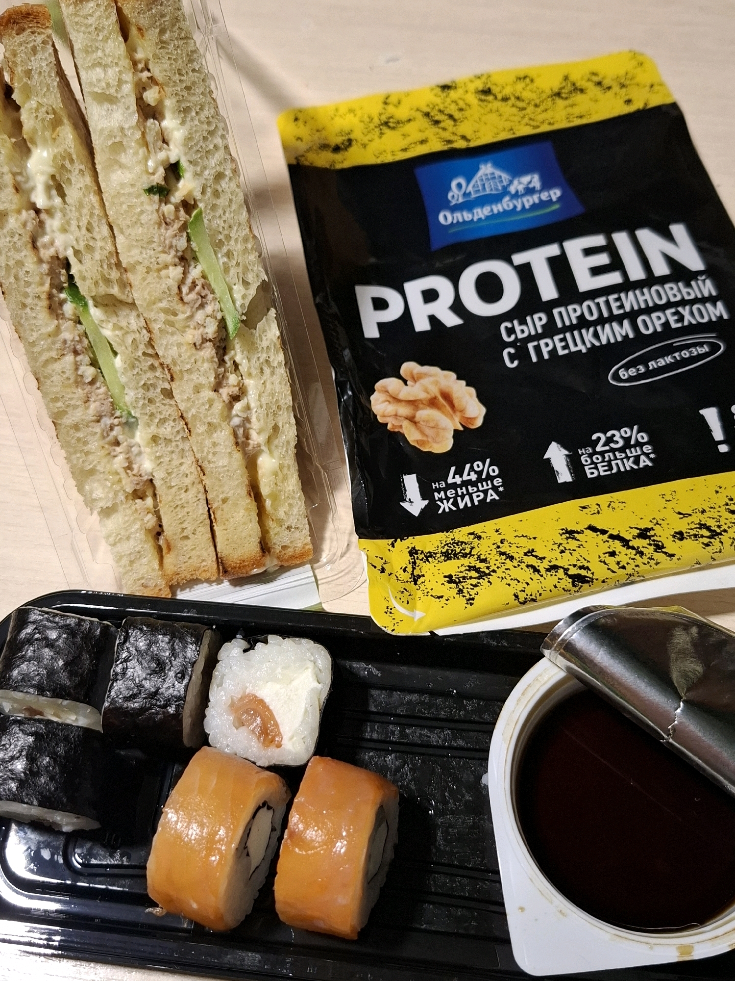 The image displays a table setting with food items, including a sandwich, sushi, and a packet of protein cheese. The sandwich is made with white bread and appears to have a filling of tuna salad and cucumber slices. The sushi consists of several rolls, some wrapped in nori seaweed and others topped with salmon. A small container of soy sauce is also present. The protein cheese packet is black with yellow accents and features text in Russian, indicating it is "Protein cheese with walnuts." The scene is indoors, with a neutral-colored tabletop visible. There are no people or animals in the image, and no specific landmarks of Tyumen, Russia are identifiable.