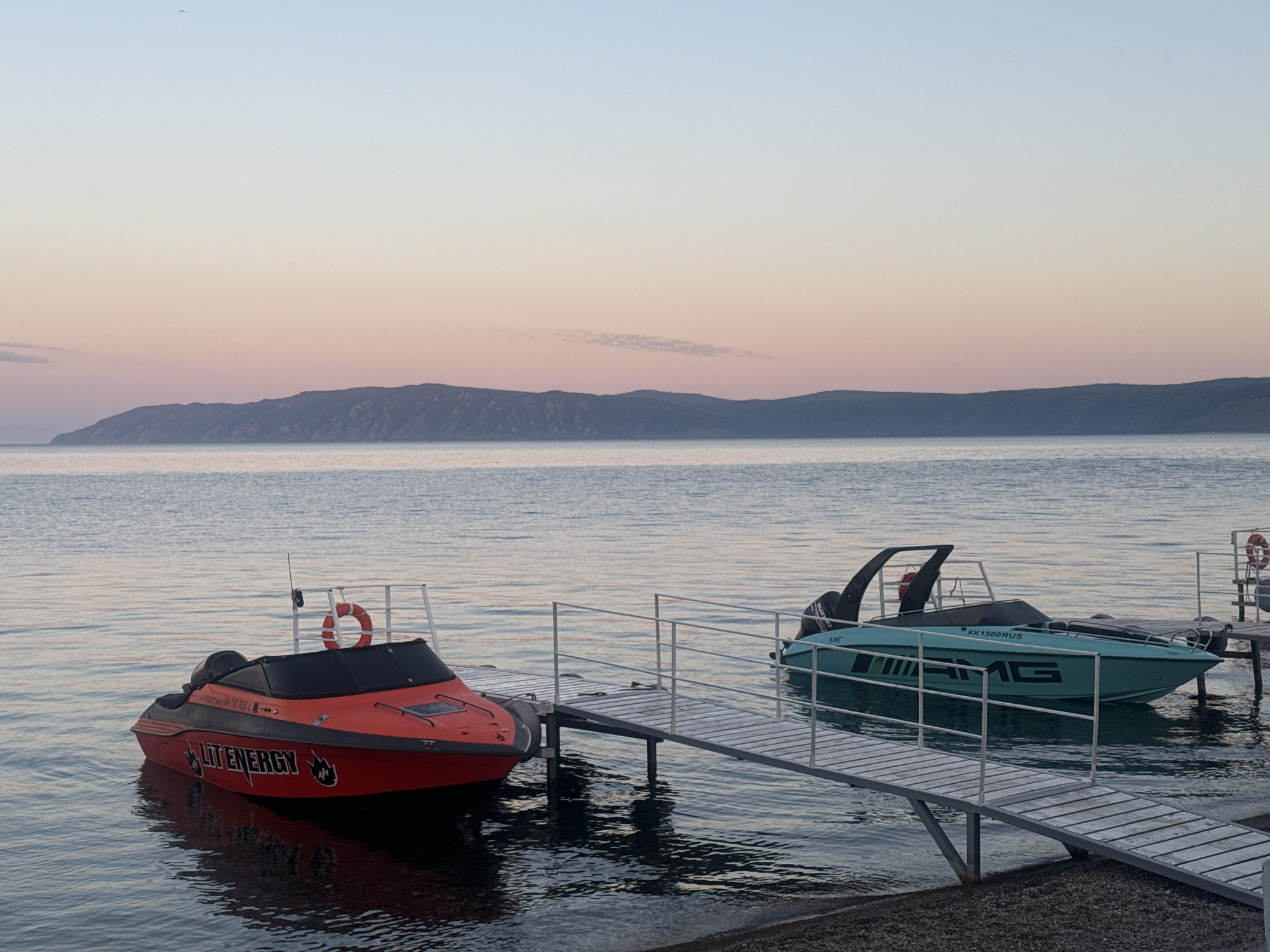 The image depicts a serene waterfront scene at sunset in Listvyanka, Russia. Two boats are moored to a wooden dock extending into calm water. The boat on the left is red with black accents and the text "LITENERGY" visible. A life preserver hangs on the railing of the dock. The boat on the right is teal with black accents and the text "AMG" prominently displayed along its side. The background features a distant, undulating mountain range under a sky painted with soft hues of pink, orange, and blue, indicative of twilight. The water reflects the sky and the boats. The shoreline in the foreground is composed of dark pebbles. There are no people visible in the image.