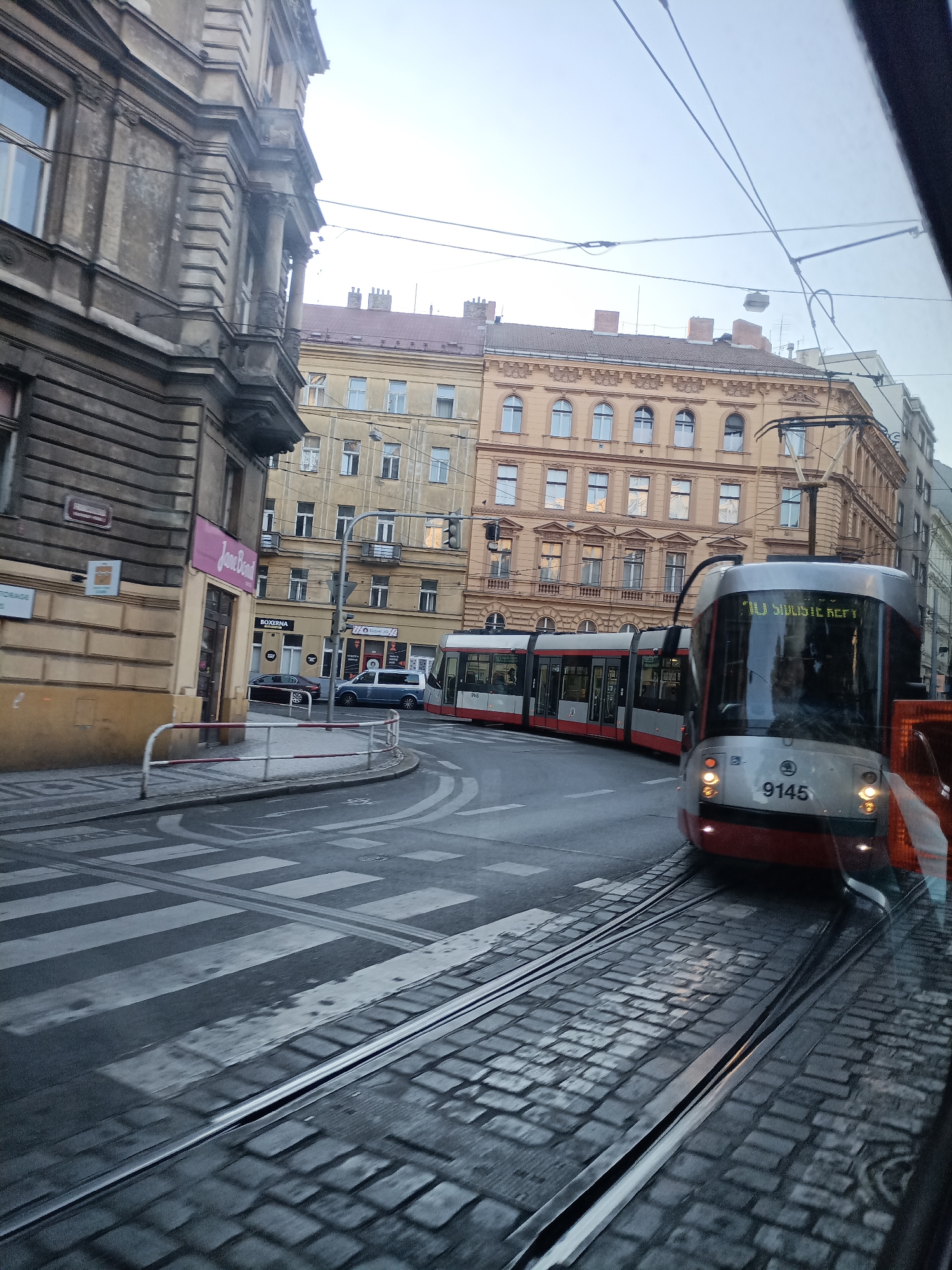 This image captures a street scene in Prague, Czechia, dominated by a modern tram moving along cobblestone tracks. The tram, numbered 9145, is white and red, with its destination displayed as "16 SUBSTETKER". It appears to be a multi-car articulated tram. In the background, ornate buildings with multiple windows and classical architectural details line the street. The sky is overcast, suggesting it is either early morning or late afternoon. The street features a pedestrian crossing marked with white stripes and tram tracks running across it. A silver van and a dark car are parked or moving on the street. The foreground shows the tram tracks and cobblestone pavement, as viewed from within another tram or vehicle. The overall impression is of a typical urban transit scene in an older European city.