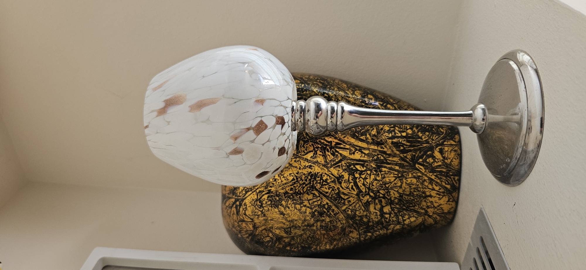 This is a still life photograph taken indoors, likely in a residential setting. The primary subject is a decorative glass candle holder with a silver base and stem, topped with a white, speckled glass shade. The shade has an irregular pattern of small, irregular shapes that appear to be copper or rose gold in color, embedded within the white glass. Behind the candle holder, there is a large, ornate vase or urn with a textured, metallic gold and black surface, featuring an intricate, almost organic pattern. The objects are placed on a light-colored surface, possibly a shelf or table, which is slightly textured. To the left of the frame, a portion of a white rectangular object, possibly a vent or part of a piece of furniture, is visible. The background is a plain, light beige wall. The lighting suggests an indoor environment with ambient light, creating soft shadows. There are no people or animals present in the image. The photograph captures a close-up view of these decorative items, emphasizing their textures and finishes. The scene is static and appears to be a curated display rather than a candid moment.