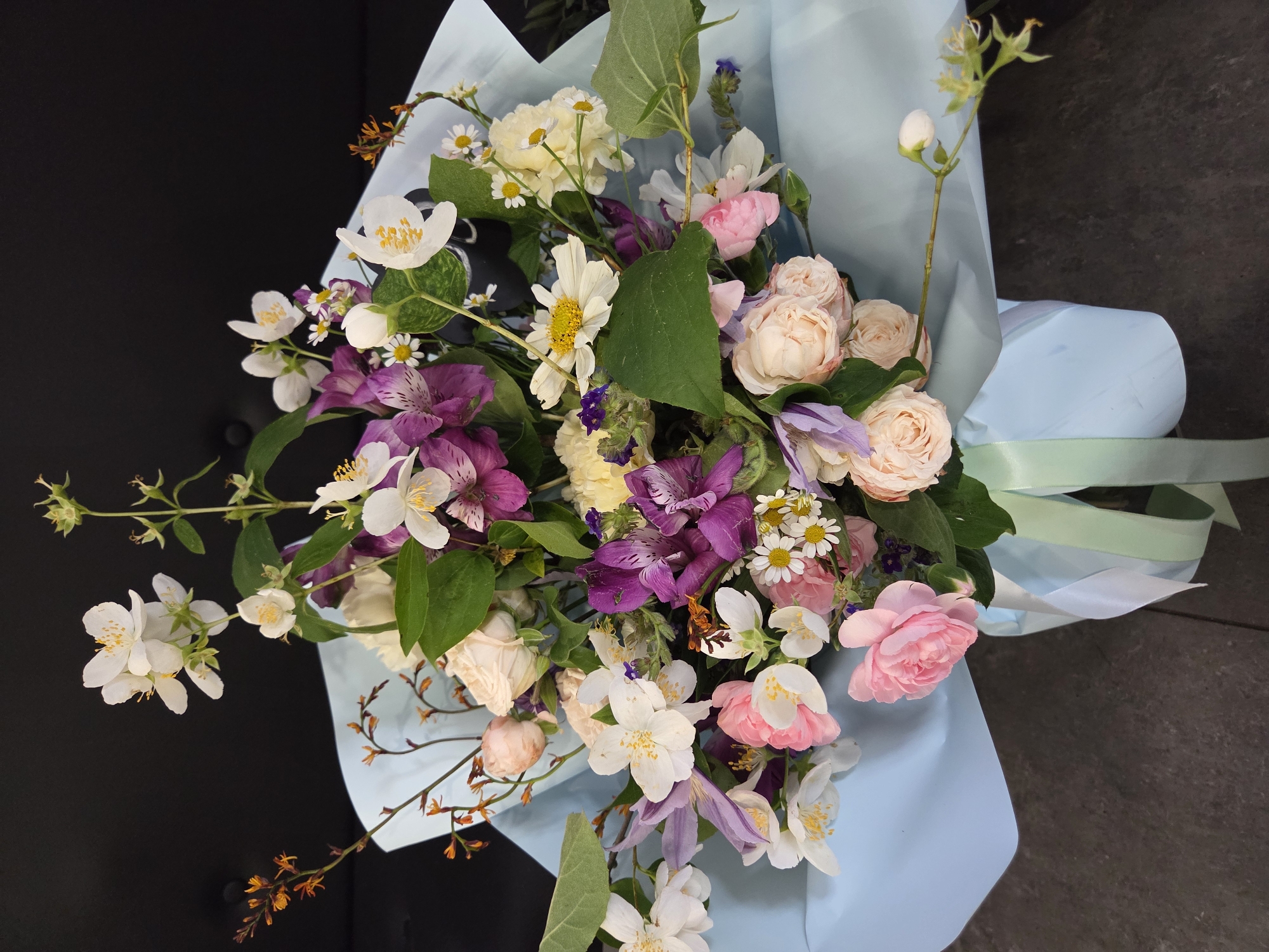 A bouquet of flowers is presented in a light blue paper wrap, tied with light green and white ribbons. The bouquet features a variety of flowers including white jasmine, pink and cream roses, purple alstroemeria, and small white daisies. The background is a dark, solid surface, likely a wall or furniture. The scene is devoid of any people or discernible location in Krakow, Poland. The lighting is even, suggesting an indoor setting.