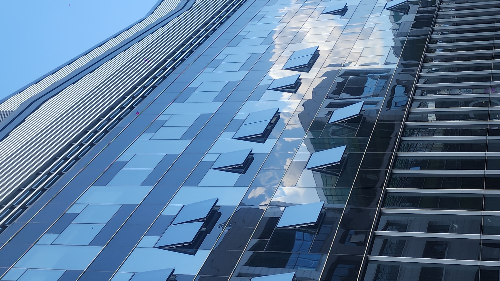 The image displays the facade of a modern building with a blue glass exterior, reflecting the sky and clouds. Several windows are partially open, angled upwards. The building features horizontal louvers along its upper section. The lower part of the facade also shows reflections of other buildings, characterized by vertical structural elements. The scene is set against a clear blue sky.