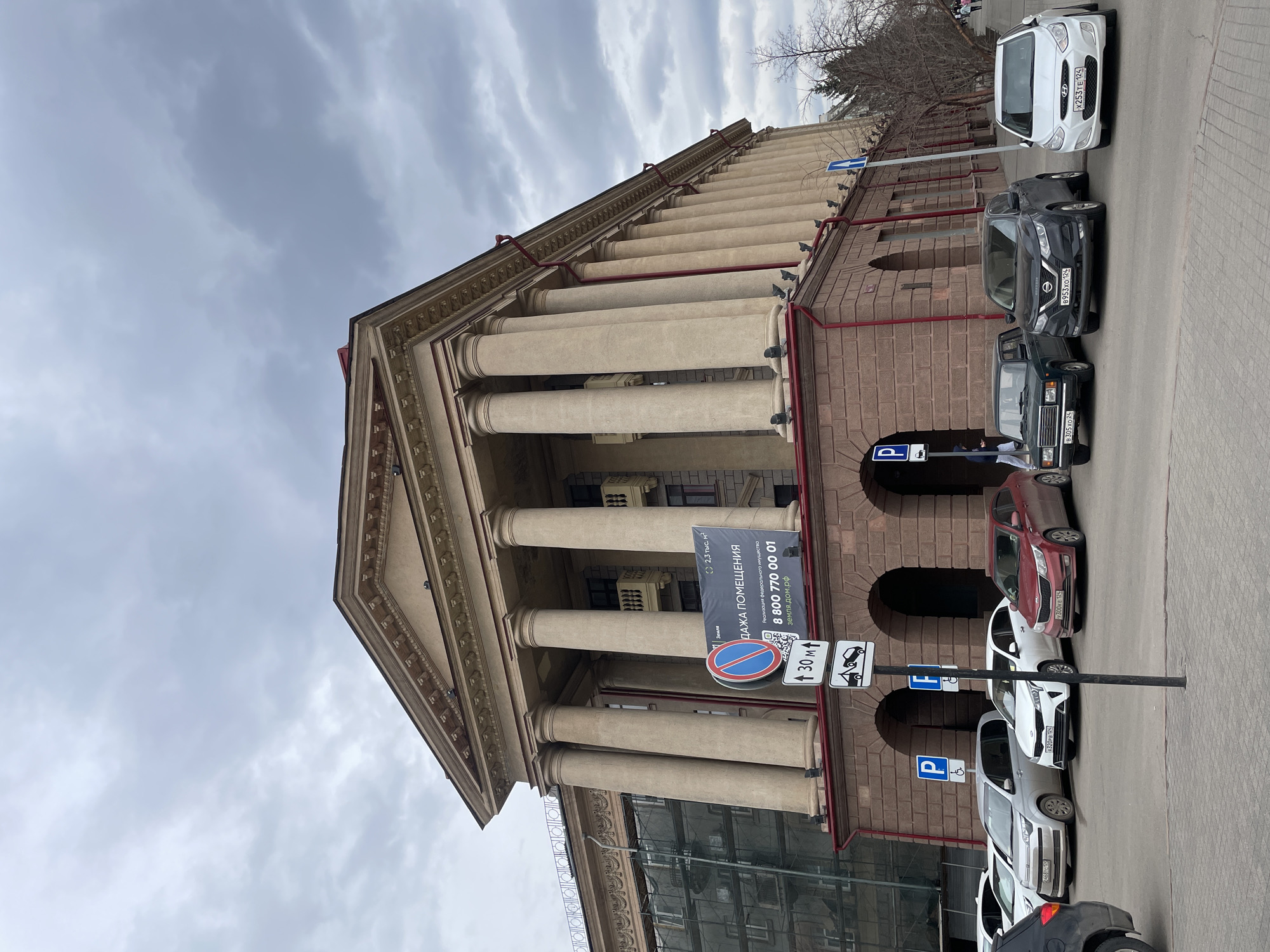 Here is a description of the image:
The image shows the facade of a building with a classical architectural style, featuring large columns. Several parked cars line the street in front of the building. There are parking signs present. There is a large advertisement sign hanging in front of the building. The sky is overcast with clouds.
Based on the number found on the advertisement sign, the building is located in Krasnoyarsk, Russia.