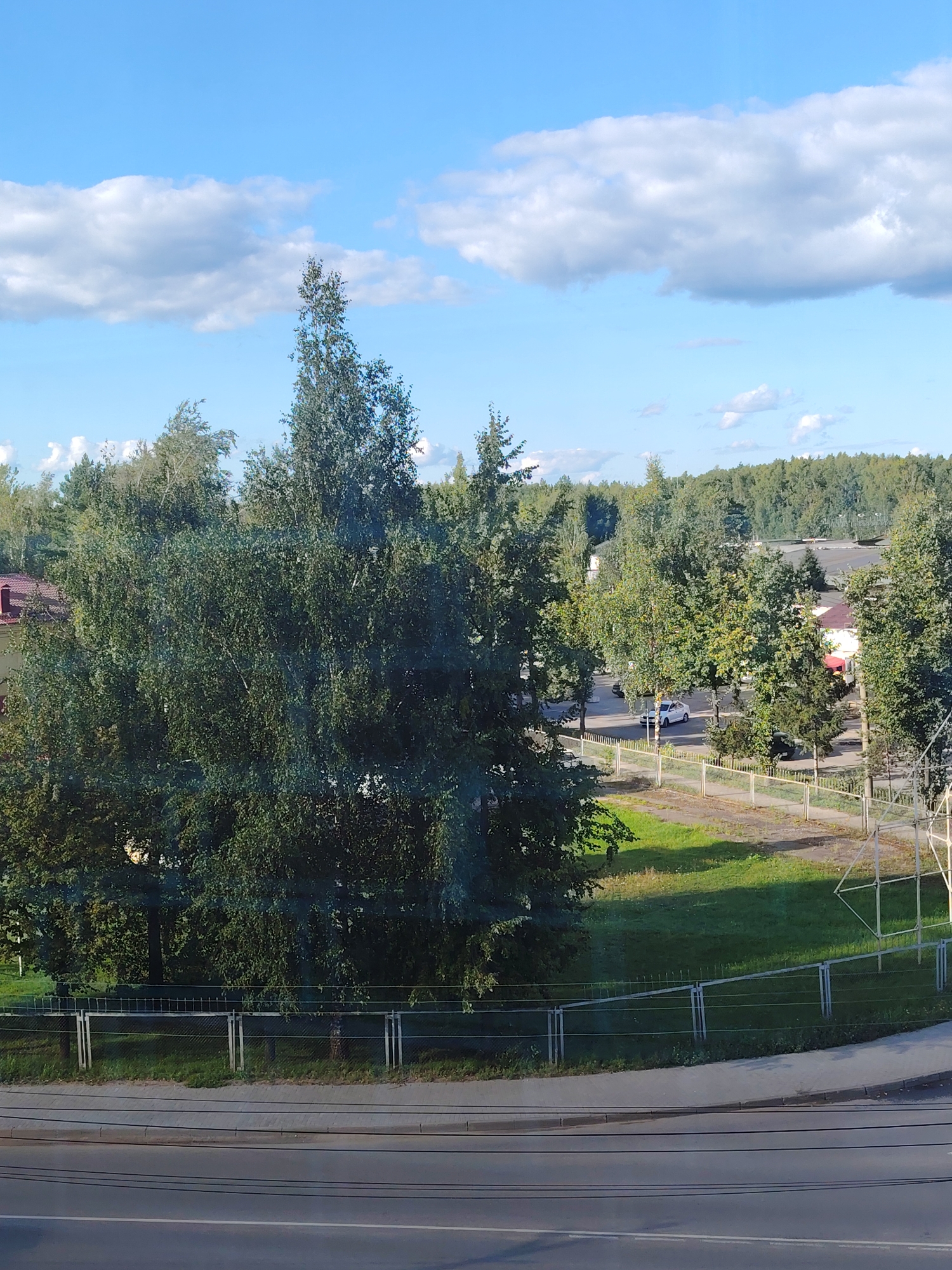 The image shows a wide-angle view of an urban and natural landscape under a partly cloudy blue sky. In the foreground, a two-lane asphalt road with power lines is visible. Adjacent to the road is a sidewalk and a grassy area bordered by a metal fence. Beyond the fence, a parking lot with a white car and a dark-colored vehicle is situated amongst trees. Further in the background, a dense forest stretches across the horizon, with some buildings, including one with a red roof, interspersed among the trees. The overall scene suggests a moderately developed area with significant green spaces, consistent with a city like Konakovo, Russia, where residential and natural environments often coexist.