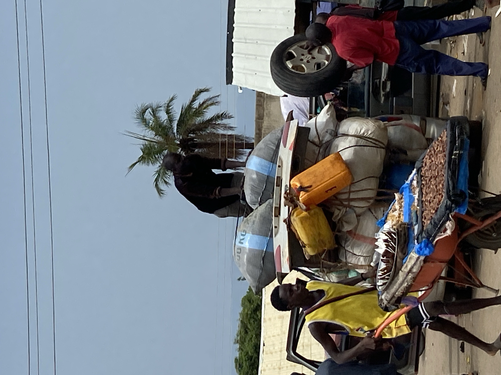 In Mararaba, Nigeria, a street scene shows a heavily loaded vehicle and several individuals. The vehicle's cargo includes large sacks, yellow containers, and items being transported by a person pulling a makeshift cart attached to it. One man in a red shirt balances a tire on top of the vehicle. Another man is standing on top of the cargo. A third man in a yellow shirt is pulling the cart alongside the vehicle. Power lines are visible in the sky above. A palm tree is partially visible in the background. Buildings are also in the background.