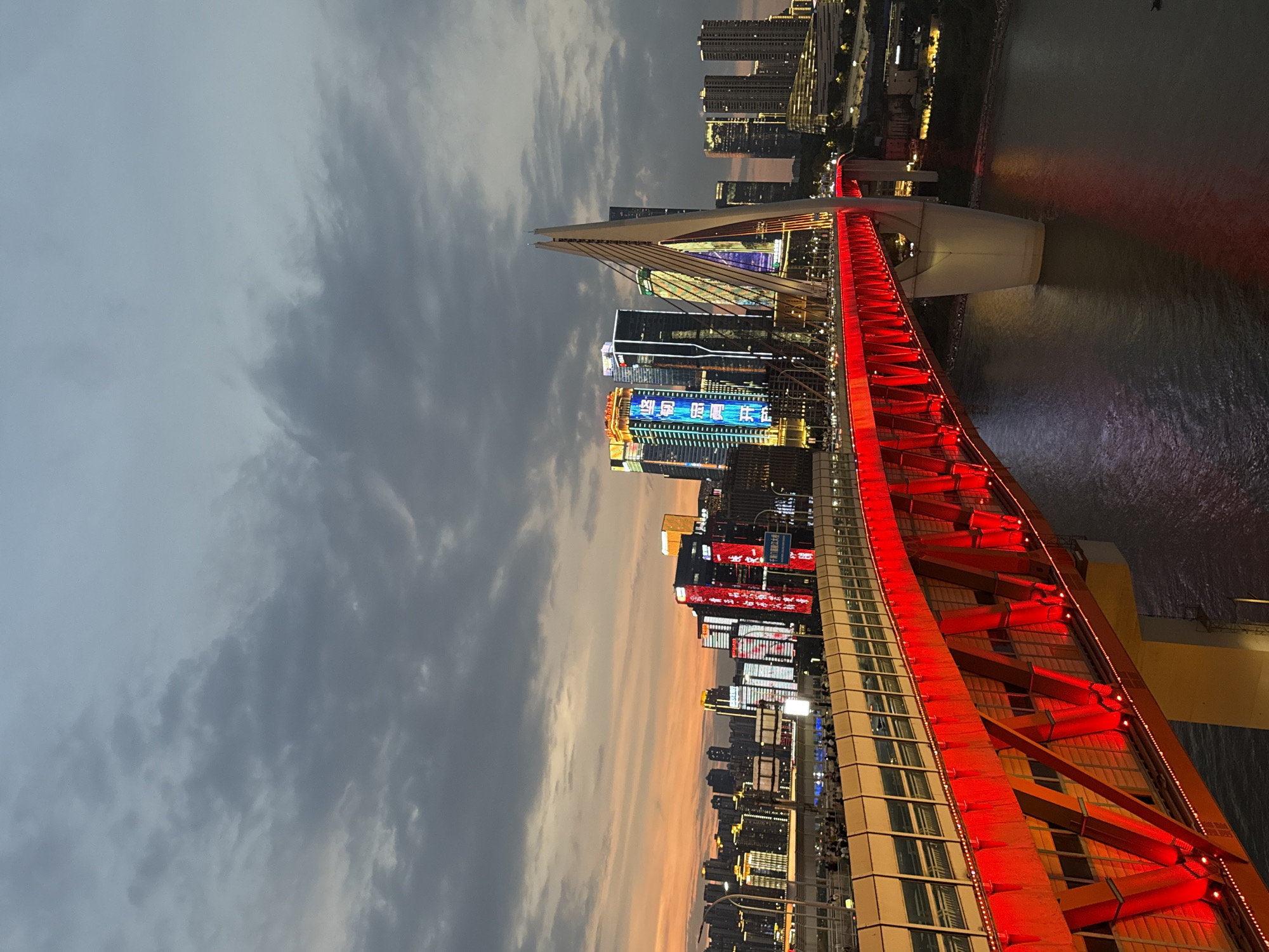 The image shows the Hongyadong Bridge at dusk in Yuzhong District, China. The bridge is illuminated with bright red lights along its structure, and the cables supporting it are white. The sky above is cloudy with a gradient of orange and gray from the setting sun. In the background, the city skyline is visible, with numerous tall buildings featuring illuminated windows and colorful neon signs. People are gathered on the pedestrian walkway of the bridge, observing the cityscape. The water of a river flows beneath the bridge, reflecting the lights of the city and the bridge itself. The overall scene captures the vibrant urban landscape of Chongqing at twilight.
