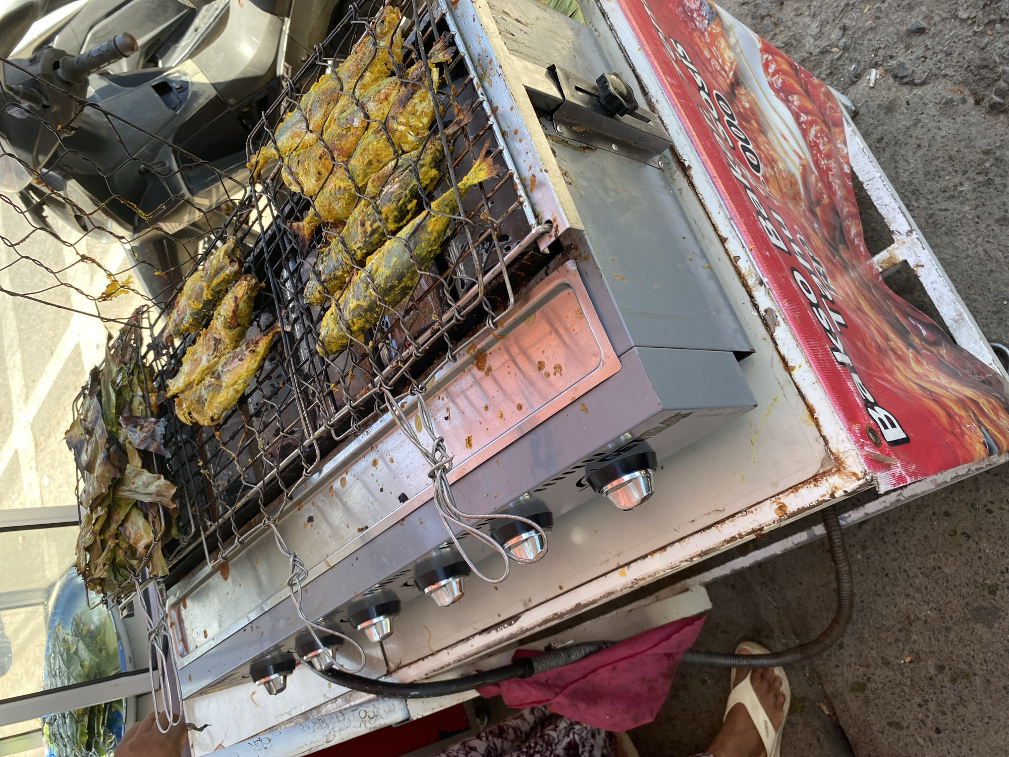 In Bayu, Indonesia, a food stall is set up outdoors, with a focus on grilled fish. A gas grill with four knobs is in the foreground, with several fish being cooked on a metal grate above it. Some of the fish are covered in a yellow spice rub. To the left of the grill, a hand is reaching towards a rack of fish wrapped in leaves, suggesting preparation for cooking. Behind the grill, a portion of a dark-colored scooter is visible, indicating the stall is likely located on a street or sidewalk. The stall itself is mounted on a wheeled cart with a vibrant red banner that has text in white and black, possibly advertising food items. The ground appears to be asphalt or concrete. A pink cloth and a person's feet wearing flip-flops are also visible to the left of the stall.