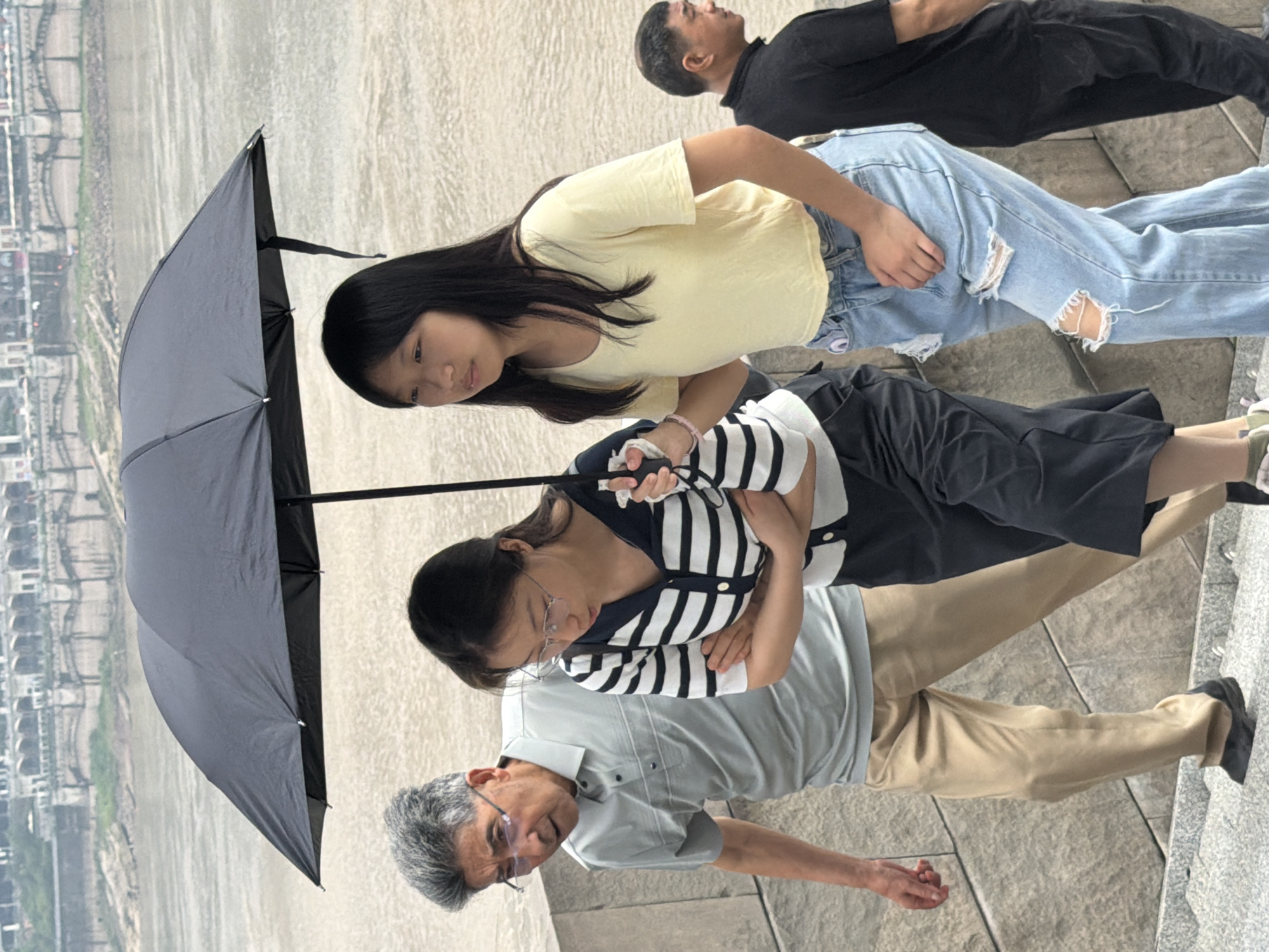 In the Yuzhong District of China, four individuals are captured walking on a stone staircase next to a body of water. A dark umbrella, held by one of the individuals, is open above three people in the foreground. An elderly man with gray hair and glasses, wearing a light gray polo shirt and tan pants, is on the left. Beside him, a young woman in a black and white striped long-sleeved shirt and dark pants is looking down. To her right, a younger woman in a yellow t-shirt and ripped blue jeans is looking forward. Further to the right and slightly behind, a man in a black t-shirt and dark pants walks away from the camera. The background shows a large body of water and a city skyline with architectural structures, suggesting an urban setting. The lighting suggests it is an overcast day.