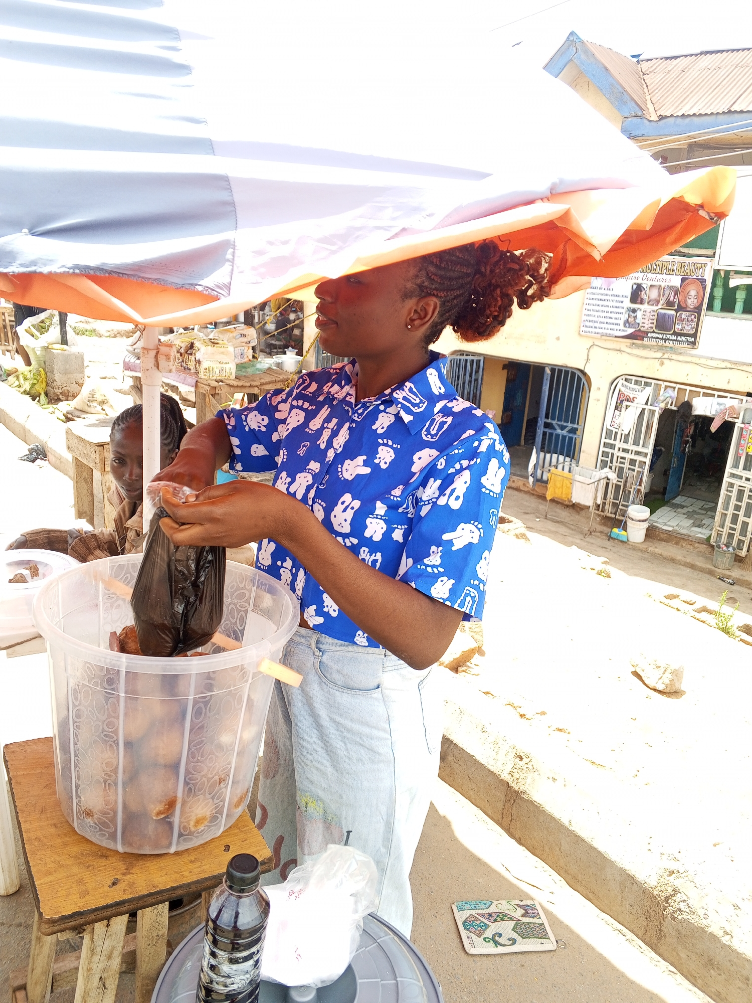 A woman stands under an orange and white umbrella in what appears to be Gwong, Nigeria, putting food into a black plastic bag. She wears a blue button-down shirt with cartoon figures and light wash jeans. A clear plastic bucket filled with what looks like food items sits on a wooden stool in front of her. A child stands to her left looking at the camera. A black bottle and white plastic bag are on a small table at the base of the stool. The street is concrete and sun-drenched. In the background, a building with blue metal doors and a sign reading "MULTIPLE BEAUTY Empire Ventures" is visible. Clothes hang outside a doorway.