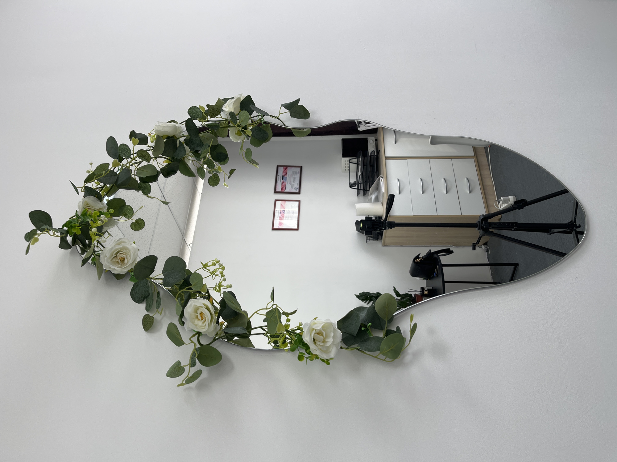 The image depicts an irregularly shaped, silver-framed mirror affixed to a plain white wall in the city of Фоки, Russia. Artificial floral garland, consisting of white roses, green eucalyptus leaves, and small green berry-like clusters, is draped around the top and left edge of the mirror. The mirror's reflection reveals an interior room with white walls and a white acoustic tile ceiling equipped with linear fluorescent lighting.
Within the reflection, two framed certificates or documents are visible on the rear wall. A light wood and white multi-drawer cabinet stands on the right, topped with office equipment including a black paper tray containing documents, a compact dark-colored safe or appliance, and an unidentified device. A black tripod, possibly for a camera or lighting, stands directly in front of this cabinet. To the left of the cabinet, a small black round table holds a black handbag and a potted green plant. The floor visible in the reflection is covered with a dark gray mat or carpet. No individuals are present in the scene.