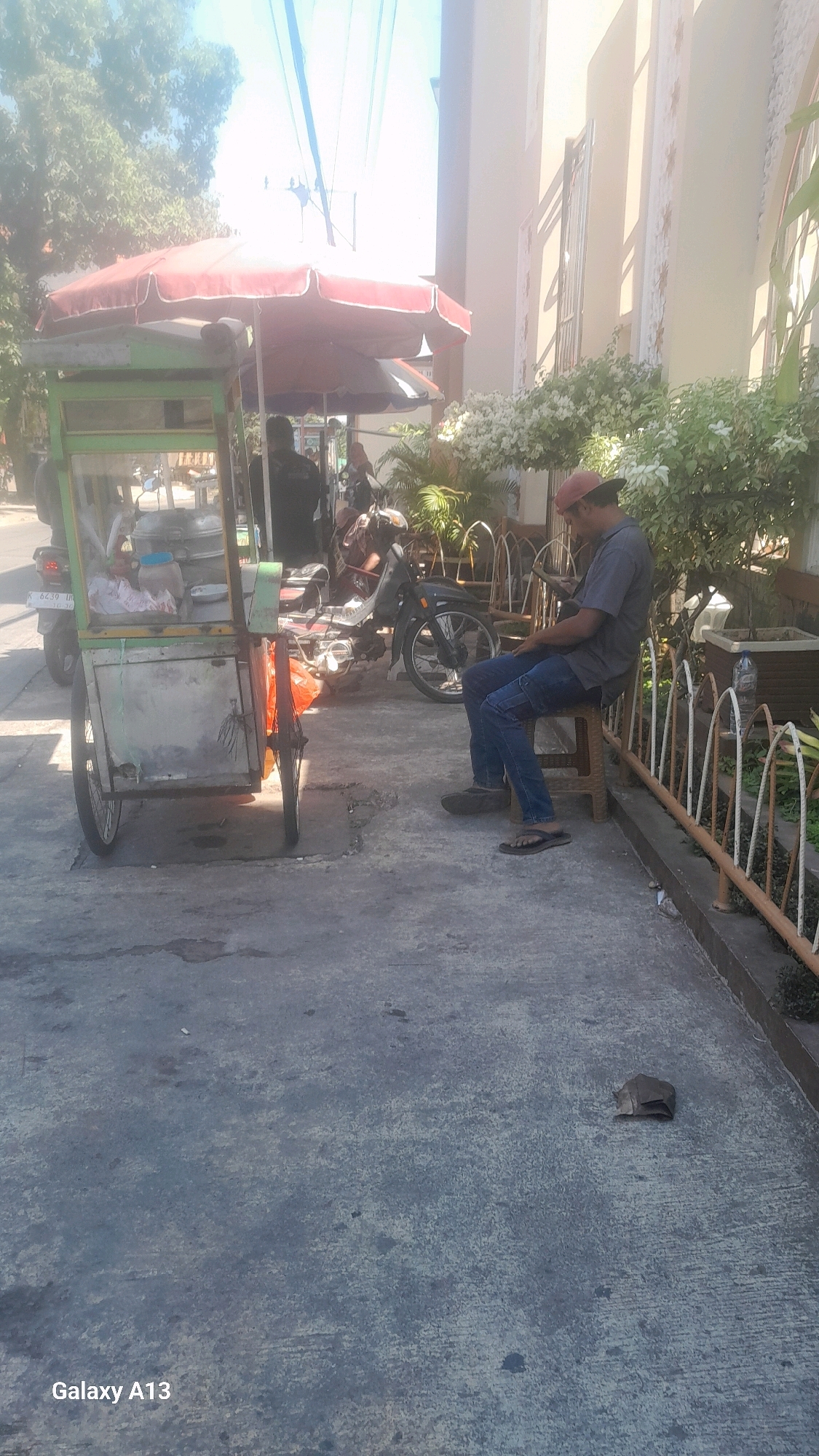 The image captures a street scene in Kirig, Indonesia, bathed in bright sunlight. In the foreground, a two-wheeled cart, likely a food or beverage vendor, is parked on a concrete pathway. The cart is covered with a red and white umbrella, and appears to be stocked with various items. To the right of the cart, a man wearing a red cap, a grey t-shirt, and blue jeans is seated on a small stool, looking down at his hands. He is wearing flip-flops. Behind him, several motorcycles are parked, and a white-flowered plant adorns the area near a building. The background reveals trees and overhead wires, suggesting an urban or semi-urban environment. The ground is a rough, grey concrete surface. A small, dark-colored object, possibly a piece of trash or a rock, rests on the ground near the seated man. The "Galaxy A13" watermark is visible in the bottom left corner of the image.