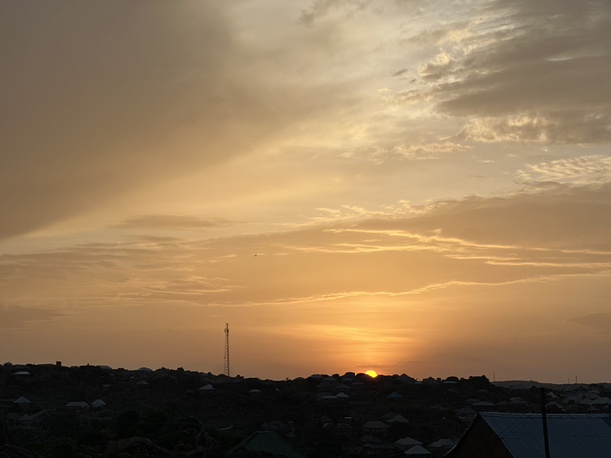 The image shows a sunset in Kabong, Nigeria. The sky is a gradient of orange and yellow, with clouds scattered throughout. The sun is partially obscured by the horizon, and the silhouettes of buildings and vegetation are visible in the foreground. A tall communication tower stands out among the buildings.