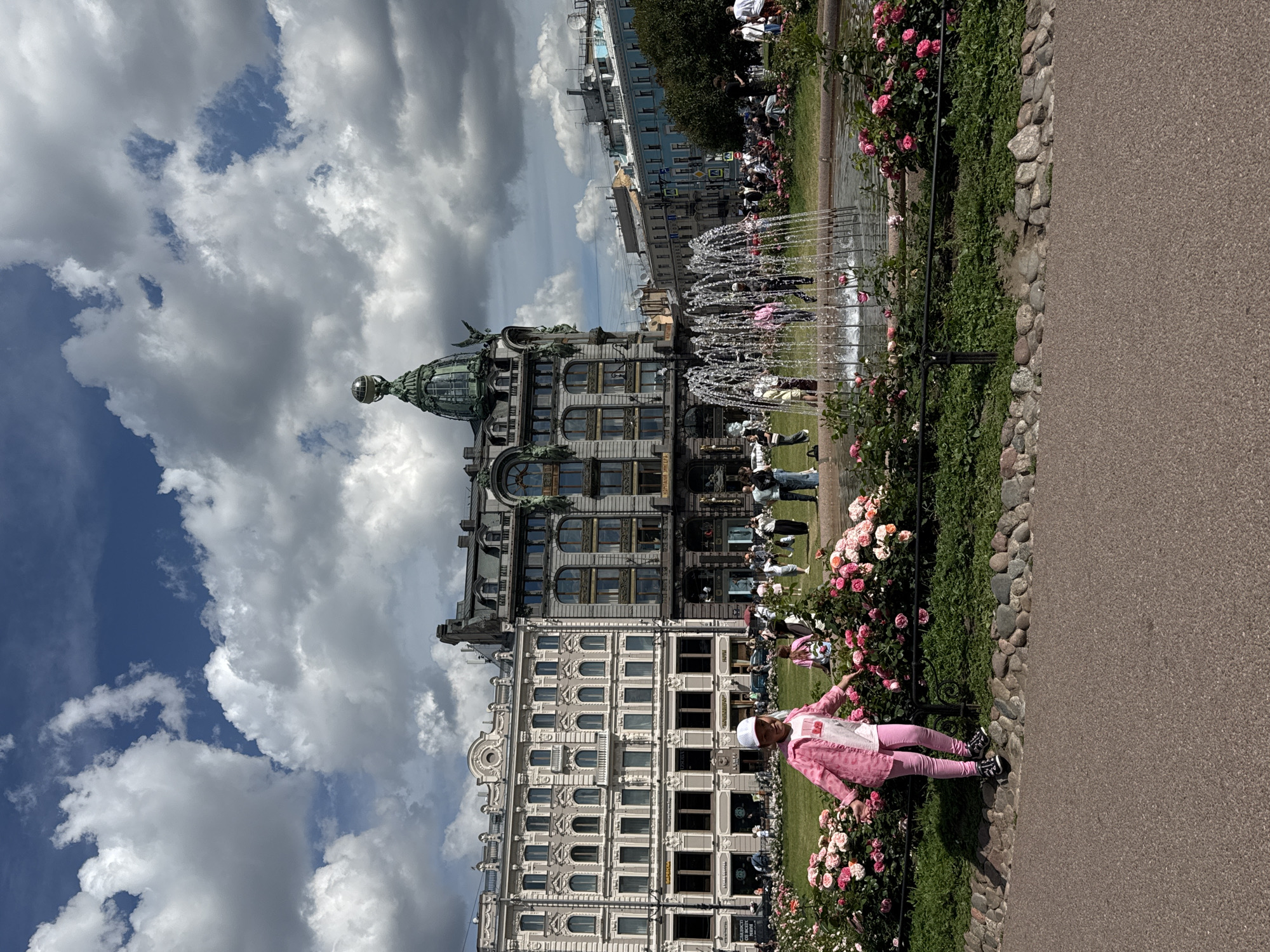 The image shows a young girl in a pink jacket and pants standing near a flower bed in a park in Saint Petersburg, Russia. In the background, there are several large, ornate buildings, a fountain spraying water, and a crowd of people. The sky is partially cloudy. The girl is looking towards the camera with her arms outstretched. The park appears to be well-maintained with lush greenery and colorful flowers. The architecture of the buildings suggests a historical and possibly European setting.
