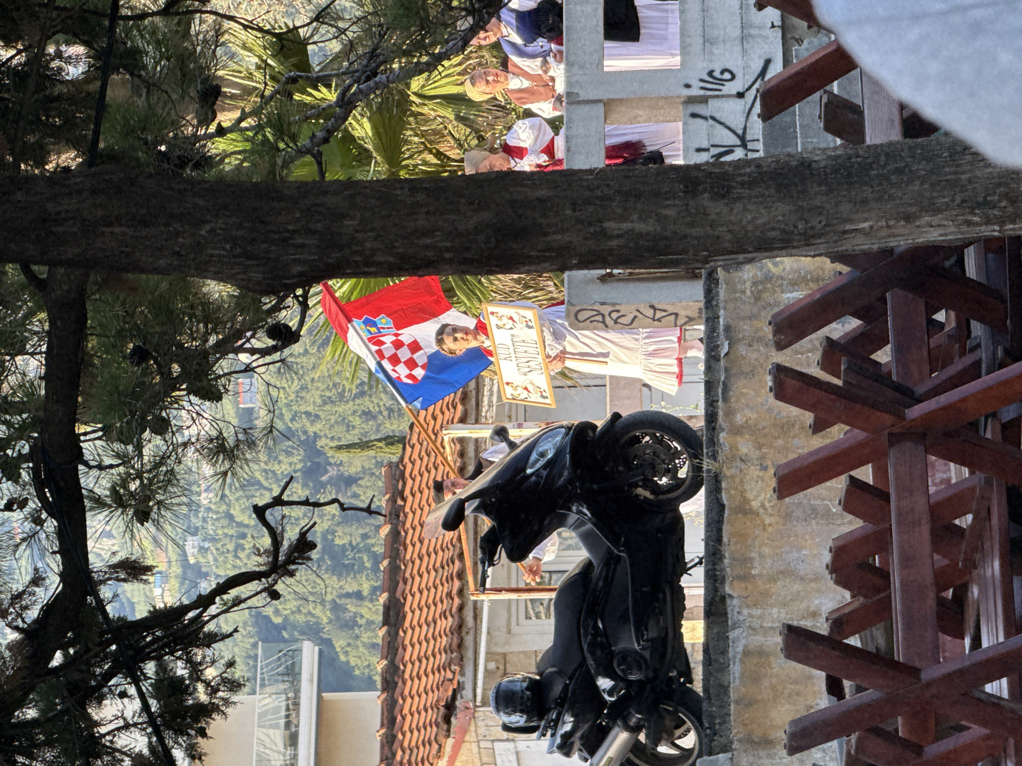 The image depicts an outdoor scene in what is identified as Budva, Montenegro. In the midground, a young woman stands on a concrete platform, facing forward, wearing a white traditional dress with red trim on the skirt and sleeves. She holds a large Croatian flag, featuring red, white, and blue horizontal stripes with a checkerboard shield and coat of arms, along with a white rectangular sign displaying "KUD 'SESVETE'" in red, stylized script. To her right, partially obscured by a thick, dark tree trunk, are several older women in various traditional Croatian garments including white blouses, vests (red, brown, and blue), and headscarves, appearing to be gathered or observing. On the left side of the platform, a black Yamaha TMAX scooter is parked, facing right, with a black helmet resting on its seat. The concrete platform's visible lower section is weathered and marked with black graffiti.
The background reveals a densely forested hillside with scattered buildings, some featuring white facades and glass balconies, suggesting a mix of natural landscape and urban development. A building with a terracotta tiled roof is visible behind the scooter. The foreground is partially framed by dark tree branches laden with pine needles and cones, particularly across the top and down the left side, and by the thick tree trunk that extends vertically from the bottom edge towards the top, obscuring parts of the scene to its right. The overall lighting suggests a bright, sunny daytime environment. In the bottom foreground, out of focus, are stacked wooden structures, possibly parts of furniture like benches or tables, characterized by their dark reddish-brown color and "X" shaped supports.