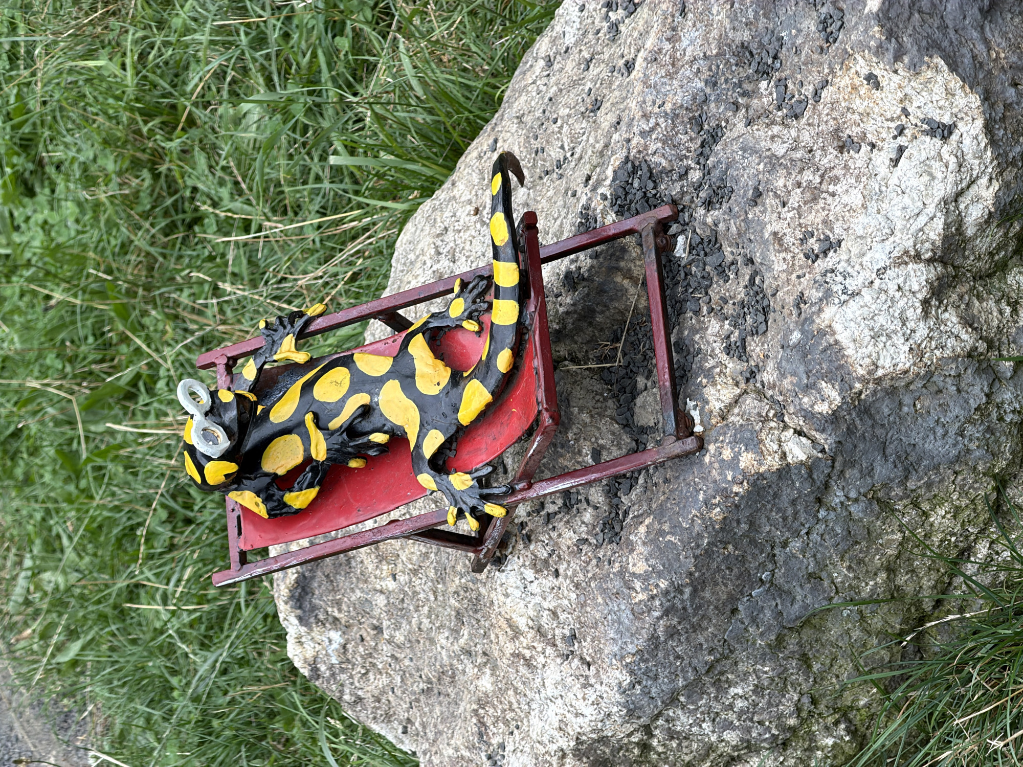 An outdoor, eye-level, full shot shows a black and yellow salamander statue wearing white goggles, resting on a red miniature deck chair. The deck chair is placed on a large, grey, rough-textured rock. Behind the rock, green grass and foliage are visible under a cloudy sky. The statue is positioned slightly off-center to the right. There are no people in the image. The overall scene conveys a sense of humorous repose. The provided context does not offer specific location information within Świeradów-Zdrój, Poland.