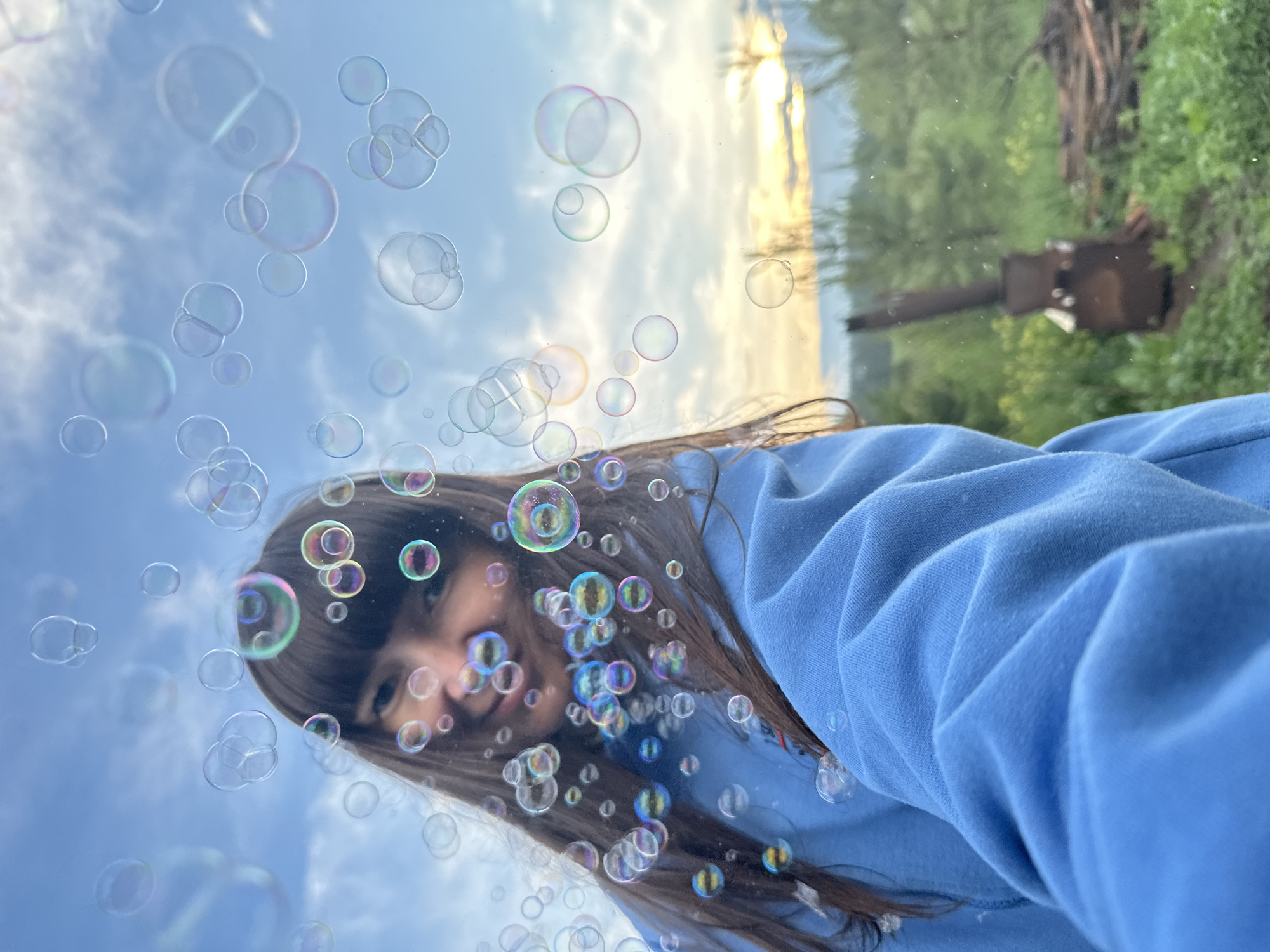 A young woman with bangs and long, dark hair is captured in a selfie, surrounded by numerous iridescent soap bubbles floating in the air. She is wearing a light blue hooded sweatshirt. The background reveals a natural outdoor setting with green foliage and trees, indicative of a rural or suburban environment. In the far background, a rustic metal structure, possibly an outdoor oven or furnace, and a pile of firewood are visible, suggesting a homestead or a backyard. The sky above is partly cloudy with hints of a sunset visible on the horizon. The overall scene evokes a sense of playful activity and a relaxed, outdoor atmosphere. The location is Buranovo, Russia.