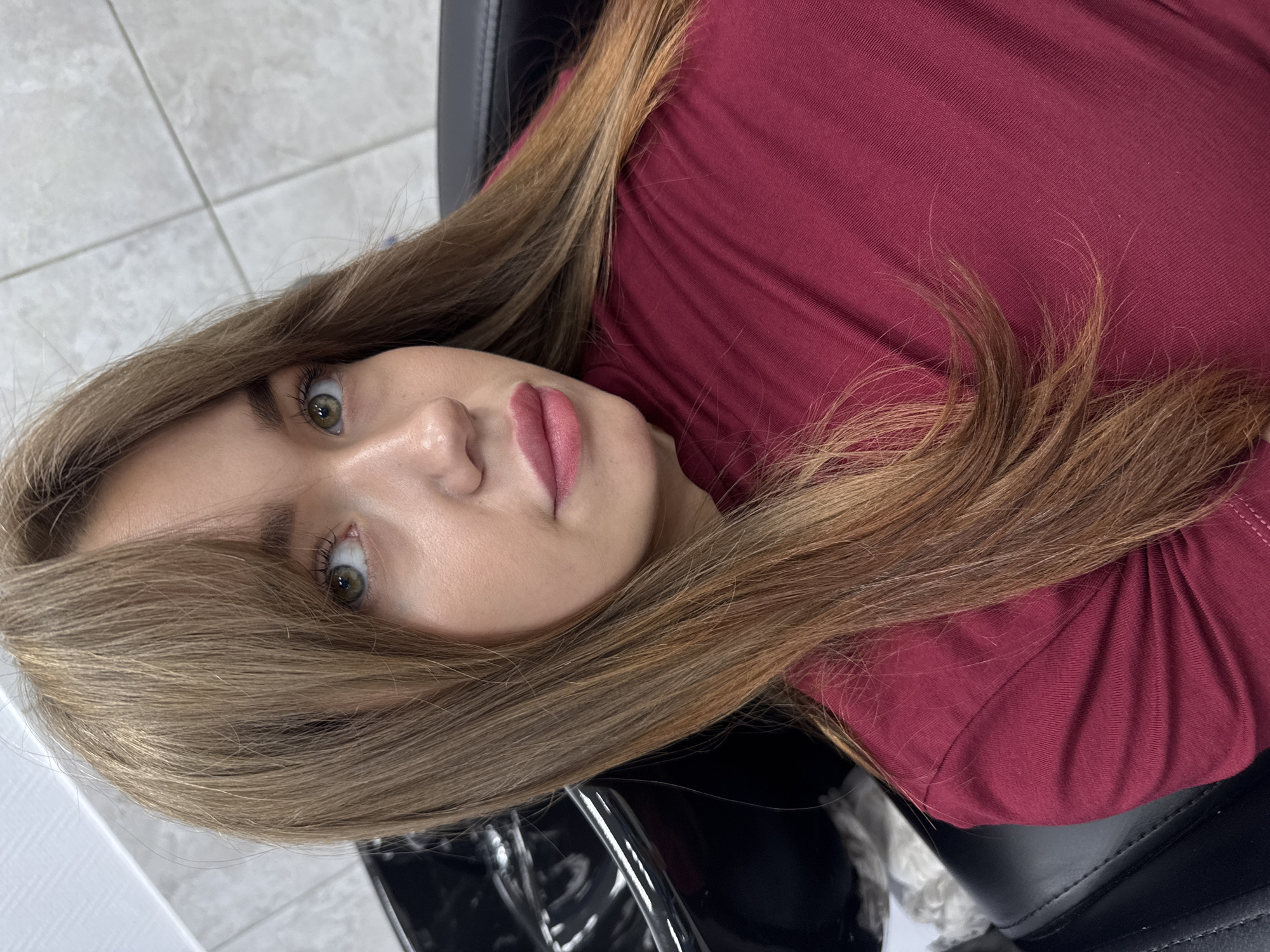 In Chaikovsky, Russia, a young woman with light brown hair and green eyes is seated in a black salon chair, looking up and to the right. She is wearing a maroon t-shirt. Her hair is long and appears to have been recently styled. To the left of the woman, a black wash basin is visible. The background shows a tiled wall.