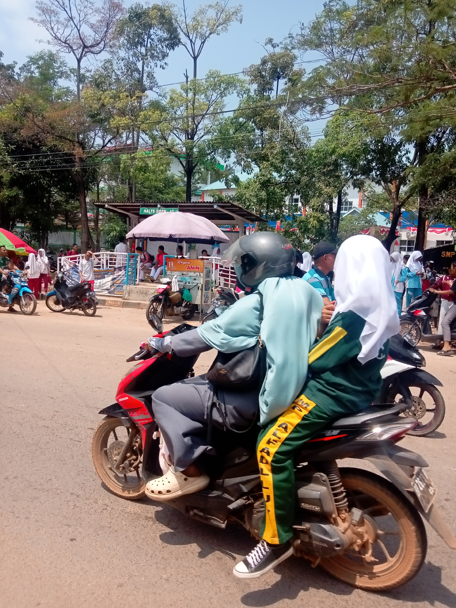 Two people are riding on a motorcycle on a paved road. The person in front is wearing a light blue long-sleeved shirt and grey pants, and a dark helmet. The person behind is wearing a white hijab and a green and yellow tracksuit. They are riding past a bus stop with a sign that says "HALTE SMPN 40". In the background, there are trees, buildings, and other people on motorcycles. The weather is sunny. The scene appears to be in Batam, Indonesia, given the presence of the SMPN 40 bus stop, which likely refers to a public junior high school in the city.