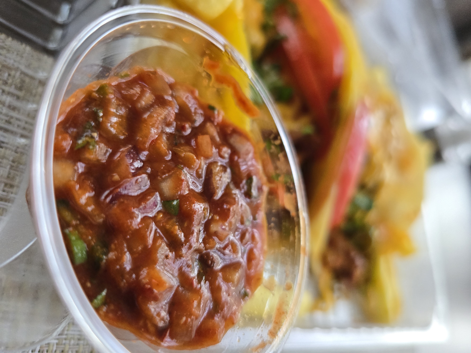 The image shows a close-up of a clear plastic container filled with a red salsa-like sauce. The salsa contains visible chunks of tomato, onion, and green herbs. In the blurred background, there appear to be tacos or other food items. The location is Unknown, Unknown.
