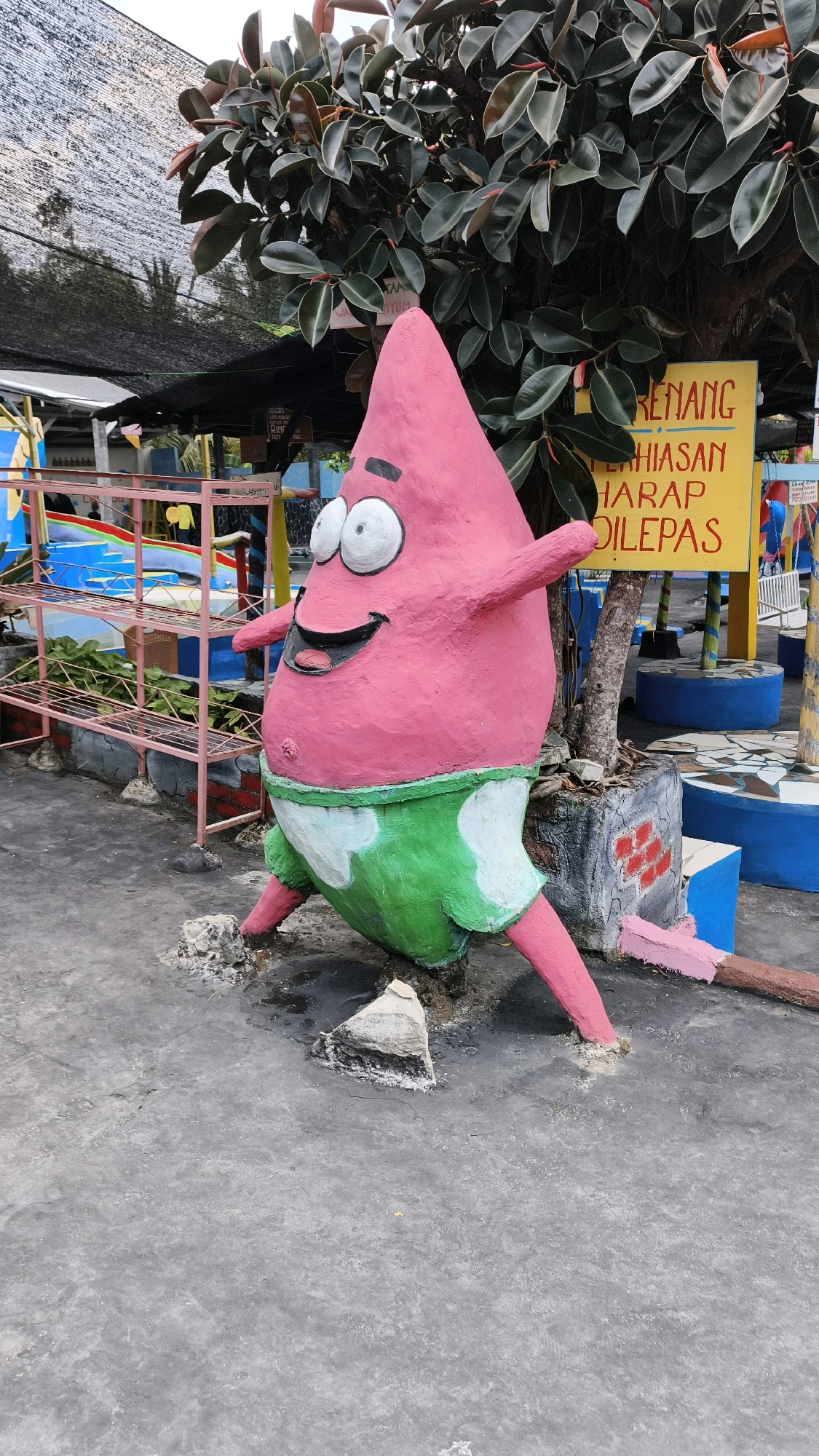 A bright pink, cartoonish statue of Patrick Star, a character from the animated series SpongeBob SquarePants, stands on a gray concrete surface in what appears to be a recreational area. The statue depicts Patrick with his signature wide eyes, open mouth, and a rotund body wearing green shorts with white wavy patterns. His limbs are outstretched, and he is positioned in a playful, slightly leaning stance.
Behind the statue, a large tree with dark, glossy leaves provides shade. A yellow sign with Indonesian text is visible behind the tree, and to the left, there are colorful playground elements, including blue structures and a pink metal shelving unit. The ground is a mottled gray concrete, with some debris and small rocks around the base of the statue. The overall impression is of an outdoor attraction, possibly a water park or amusement center, in a tropical or subtropical climate given the lush foliage. The location is identified as Air Paoh, Indonesia.