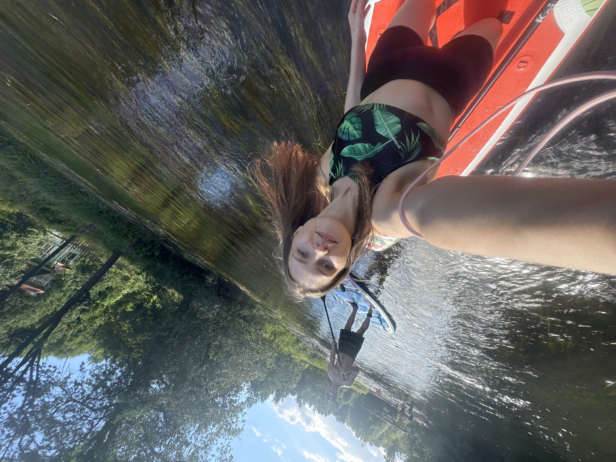 In Старосиверская, Russia, on a sunny day, a woman is taking a selfie while paddleboarding on a river. She is wearing a black bikini top with a green leaf pattern and black shorts. Her red paddleboard is in the foreground, and she is holding a paddle. In the background, a man is paddleboarding on another board, wearing dark shorts. The river is surrounded by lush green trees and vegetation. The water reflects the sky and sunlight, creating a shimmering effect. The scene appears to be a recreational outing on a warm day.