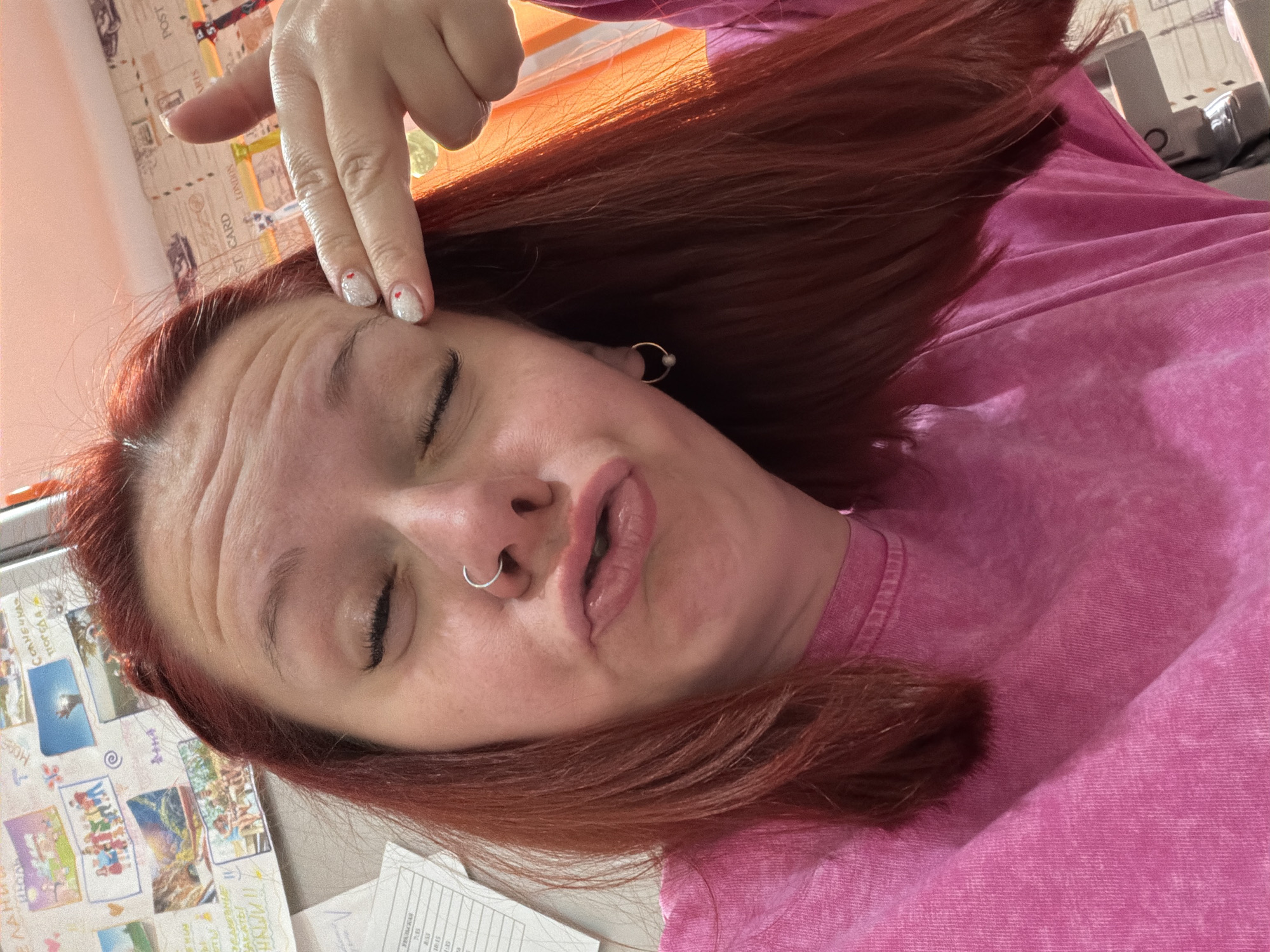 A selfie shows a fair-skinned person with auburn hair wearing a pink shirt. The person's eyes are closed, and they are making a face with their lips pursed. Their fingers touch their forehead. A nose ring is visible, as well as an earring. A window is visible behind the person, and a bulletin board with images on it can be seen, along with white papers. The location is possibly in Chaikovsky, Russia, based on the bulletin board's contents.