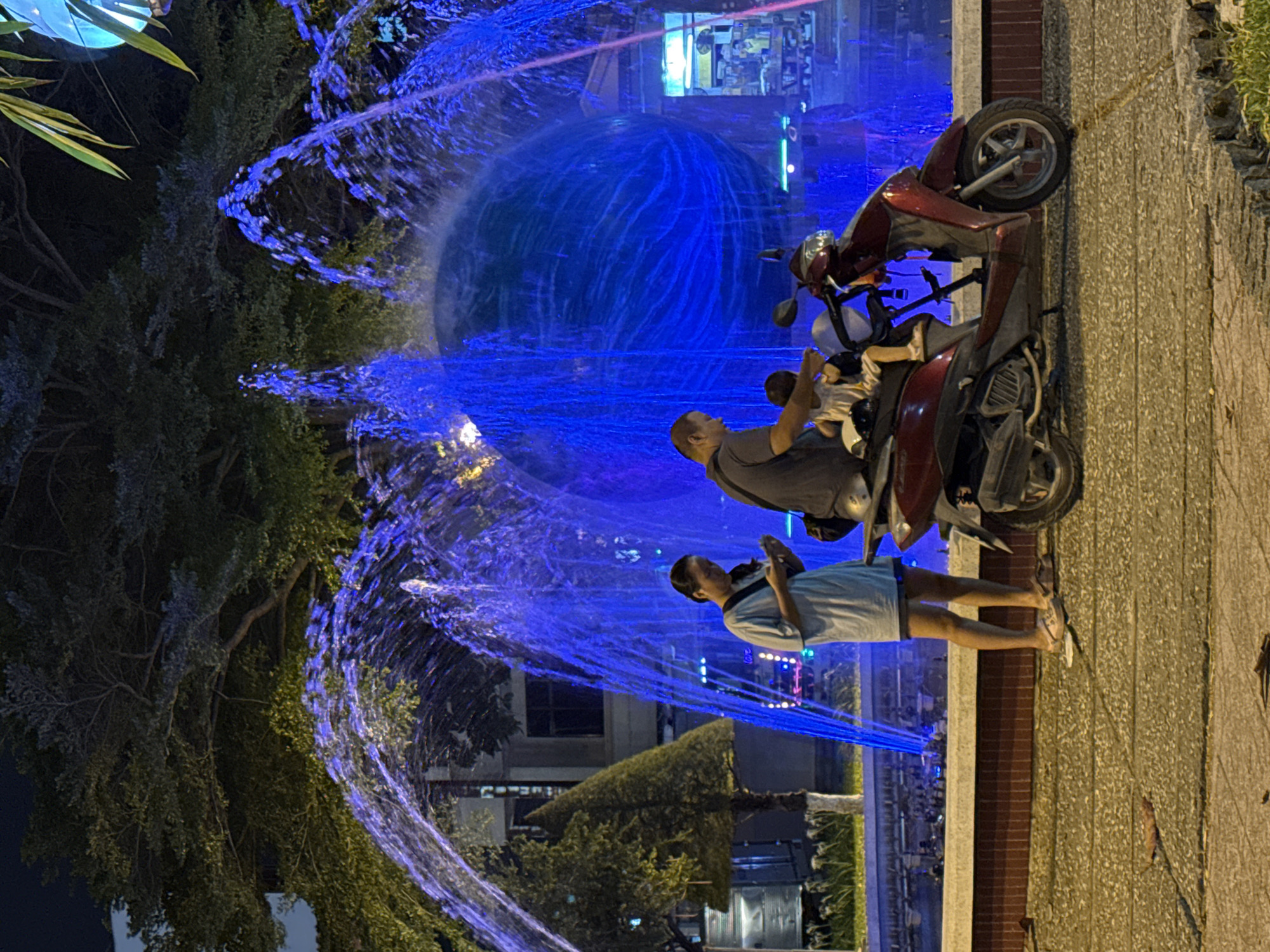The image depicts a nighttime scene in Nha Trang Ward, Vietnam, featuring a brightly lit fountain and several people interacting with a scooter. The fountain, illuminated with vibrant blue lights, forms the centerpiece of the scene, with water jets creating a dynamic visual effect.
In the foreground, a man is seated on a red scooter, holding a young child on his lap. He appears to be looking towards the fountain. Next to him, a young woman in a light blue dress stands on the sidewalk, looking in the same direction. Her posture suggests she might be observing the fountain or waiting.
The scooter is parked on a textured sidewalk next to a low brick wall, beyond which is the fountain's watery display and a glimpse of buildings in the background. Trees with lush foliage are visible on the left side of the image, adding a natural element to the urban setting. The lighting from the fountain and surrounding areas suggests it is nighttime. The overall atmosphere seems relaxed and perhaps a moment of shared enjoyment of the illuminated fountain. There is no visible text in the image.