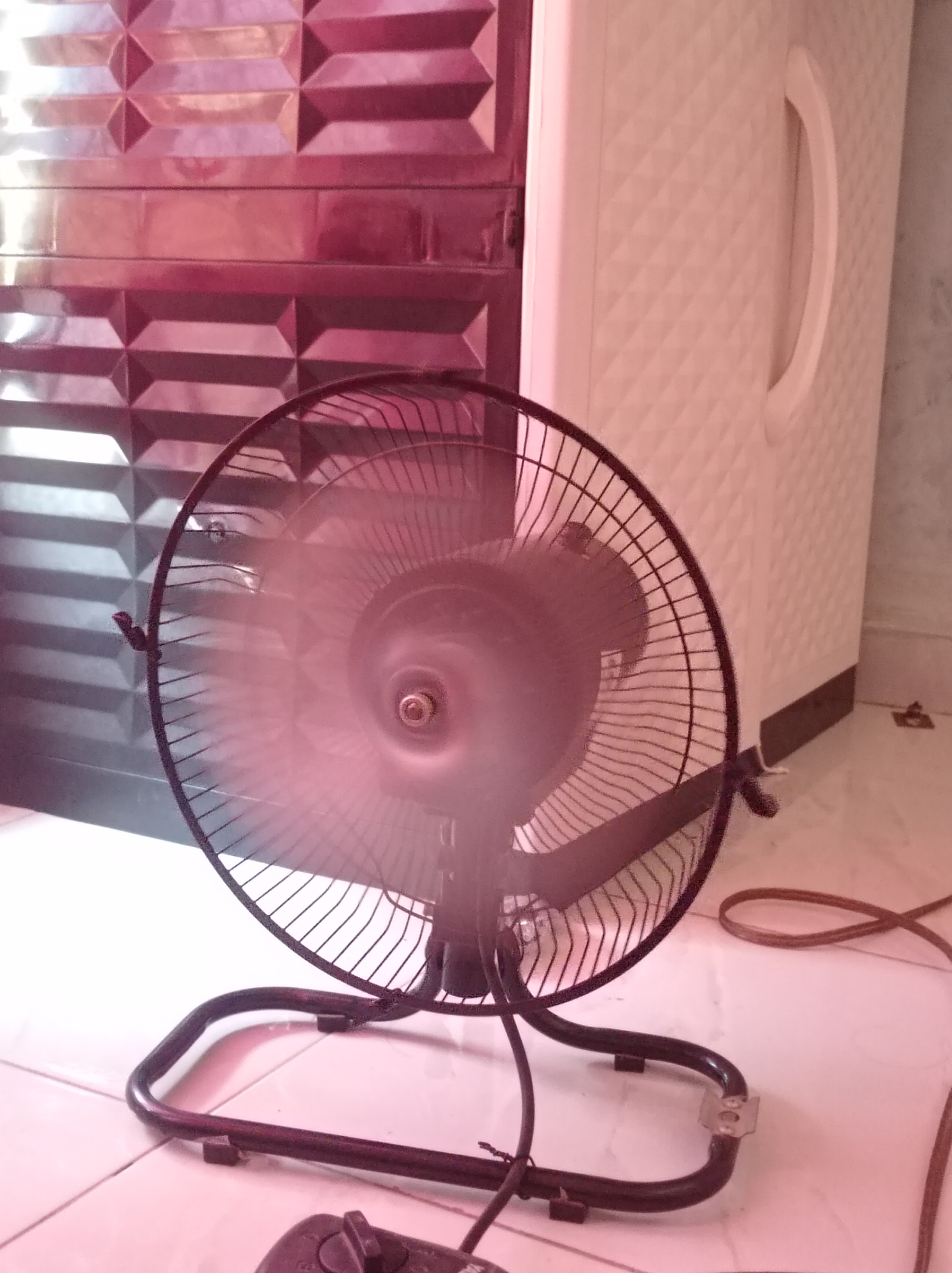 A black oscillating fan with blurred blades is positioned on a tiled floor. Behind the fan, a dark maroon, textured wall is visible, and to the right, a tall, white plastic cabinet with a diamond pattern and a curved handle stands. The fan is connected to a brown electrical cord that snakes across the floor. The scene appears to be indoors, likely in a residential setting in Sumbernadi, Indonesia, given the tiled flooring and common household items.